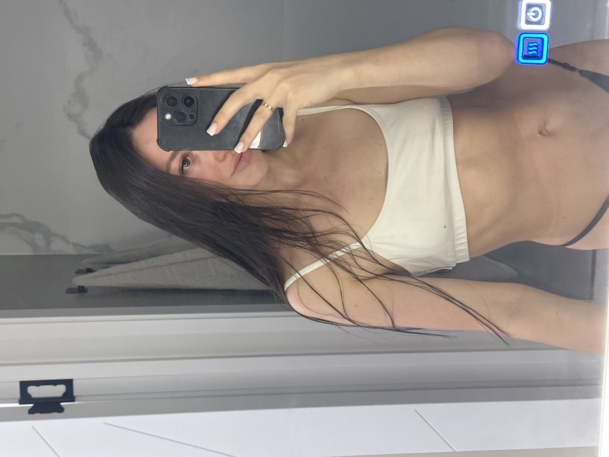 In a bathroom, a young woman with long, dark hair takes a mirror selfie. She is wearing a white bikini top and dark bikini bottoms. Her phone is held up, obscuring part of her face. The bathroom features a gray marble-patterned wall and white doors. There are towels hanging on hooks on the wall, and an illuminated control panel with blue icons is visible on the right side of the mirror. The scene is captured from the chest up, with a focus on the woman and her reflection.