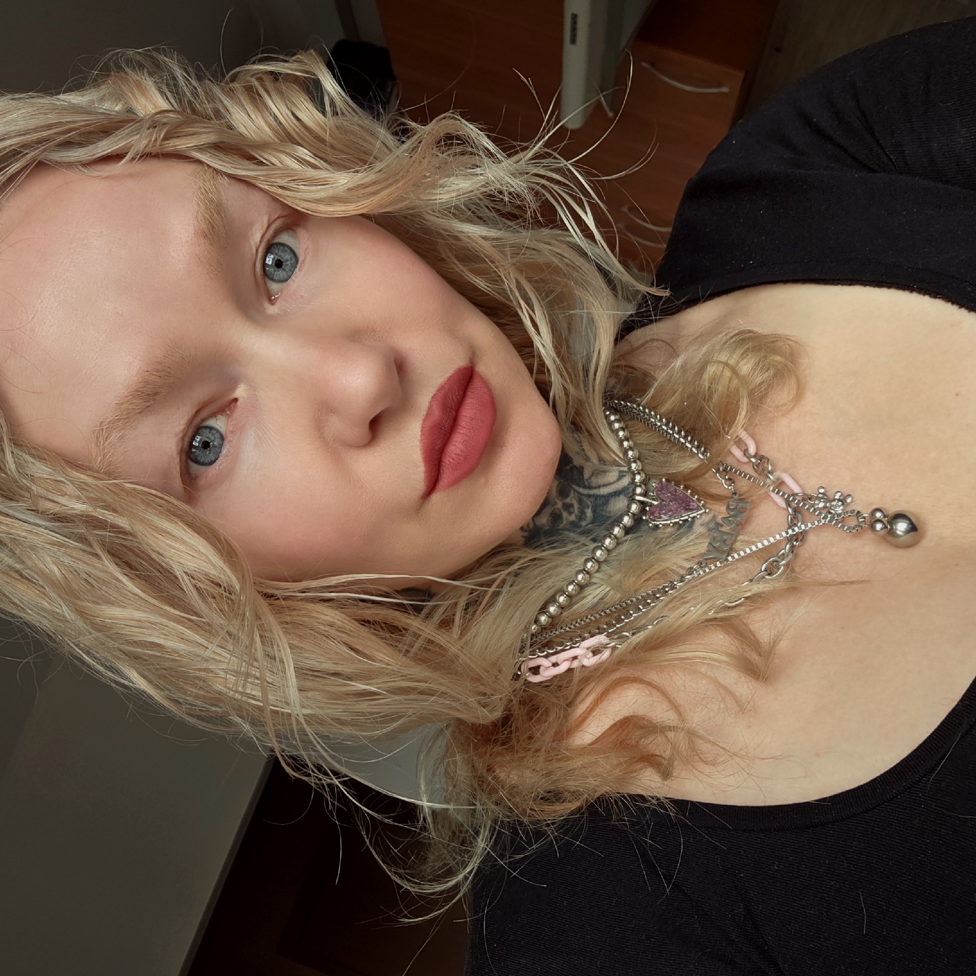 A pale-skinned young woman with light blonde, curly hair is the primary subject. She has blue eyes and wears red lipstick. She is wearing a black top, and several necklaces are visible around her neck, including one with a pink heart-shaped pendant and another with silver-colored beads. A tattoo is partially visible on her neck. The background suggests an indoor setting. The location is not discernible.