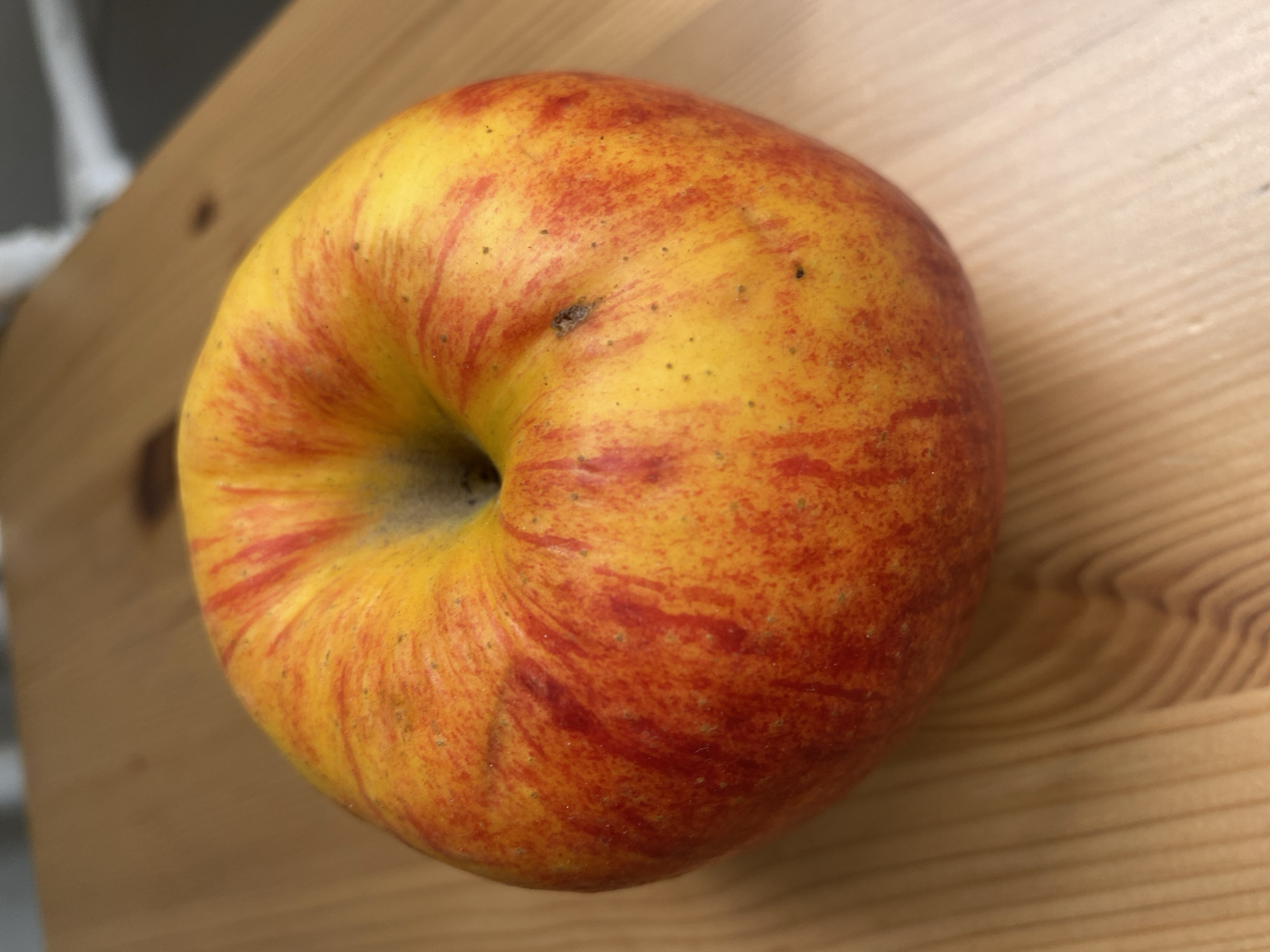 A top-down, close-up shot shows a yellow and red apple on a light brown wooden surface with visible grain patterns. The apple has a deep dimple at the top. The surface appears to be a table. The location is not discernible from the image.
