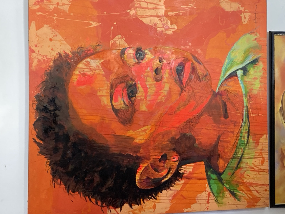 The image displays a large, vertically oriented expressionistic painting, located in the city of Ikate, Nigeria. The artwork portrays the head and upper torso of a child with dark, curly hair, gazing upwards and to the right with a slightly open mouth. The child's left ear features a small, red circular earring. The predominant color palette consists of vivid orange and red hues, characterized by prominent brushstrokes, drips, and splatters that give the painting a textured appearance. A green garment is visible on the child's upper body. The painting is affixed to a plain white wall. A portion of a second framed artwork, encased in a dark frame, is visible along the bottom edge of the image.