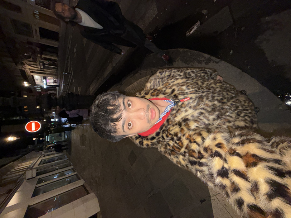 A man in a leopard-print coat takes a selfie outdoors at night. He wears a red collared shirt underneath. Behind him, another person in a dark jacket walks away. A building with lighted windows, a street sign, and pedestrians are in the background, captured with a blurry, rotating effect. The ground is dark, and a hint of light suggests a distant city.