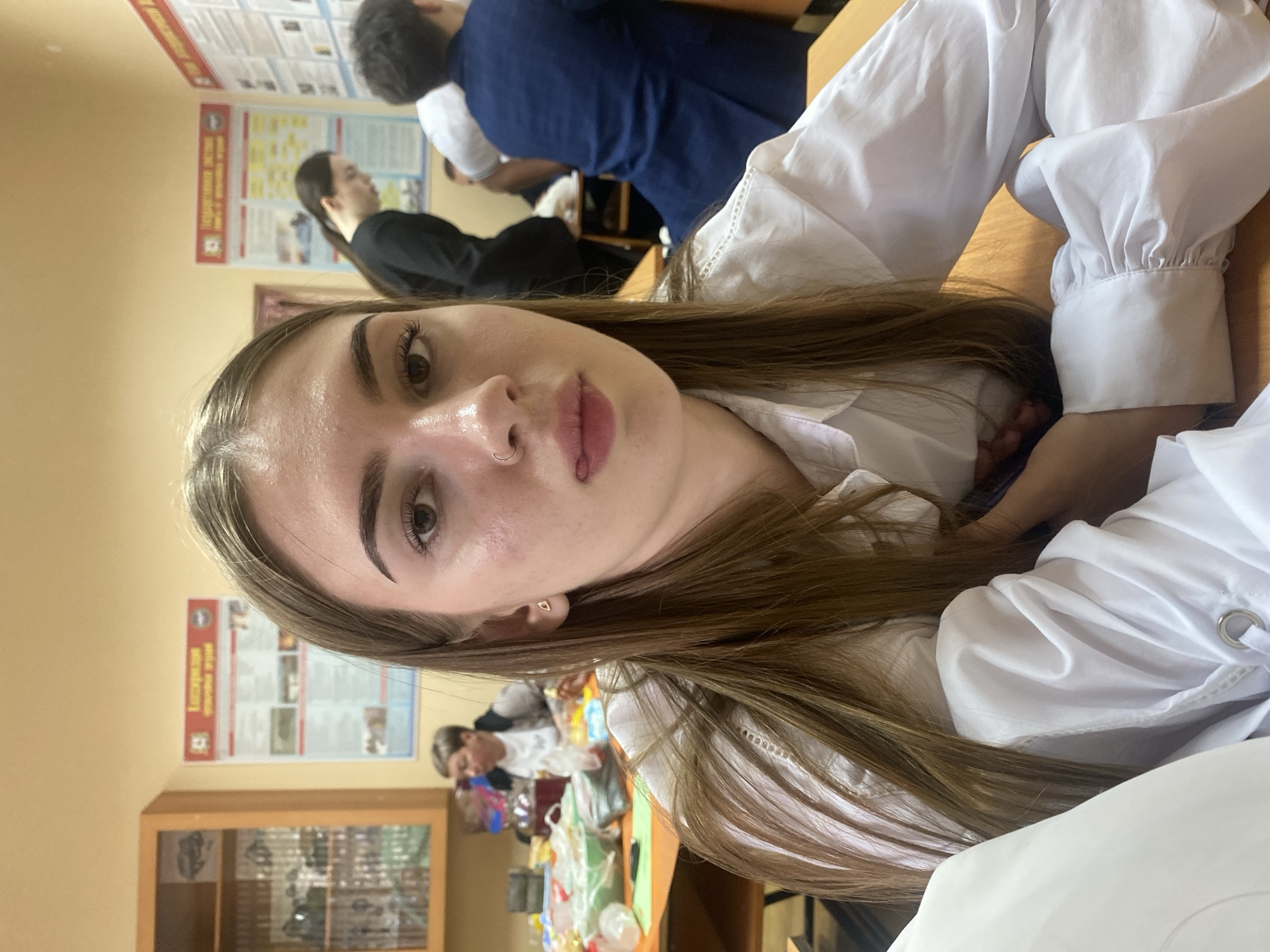 The image shows a young woman in the foreground, seated at a wooden desk. She has long, light brown hair and is wearing a white collared shirt with ruffles at the cuffs. Her face is turned towards the camera, and she is sticking her tongue out slightly. She is wearing a small gold hoop earring in her left ear and a nose ring. In the background, other people are visible, also seated at desks. Some are looking forward, while others are engaged in conversation. Posters with text and images are displayed on the walls behind them. The environment appears to be a classroom or a similar educational setting. There are some supplies on a table in the midground, including what appear to be food items and containers. The lighting suggests an indoor setting with overhead lights. The overall impression is a candid snapshot taken within a school or university.