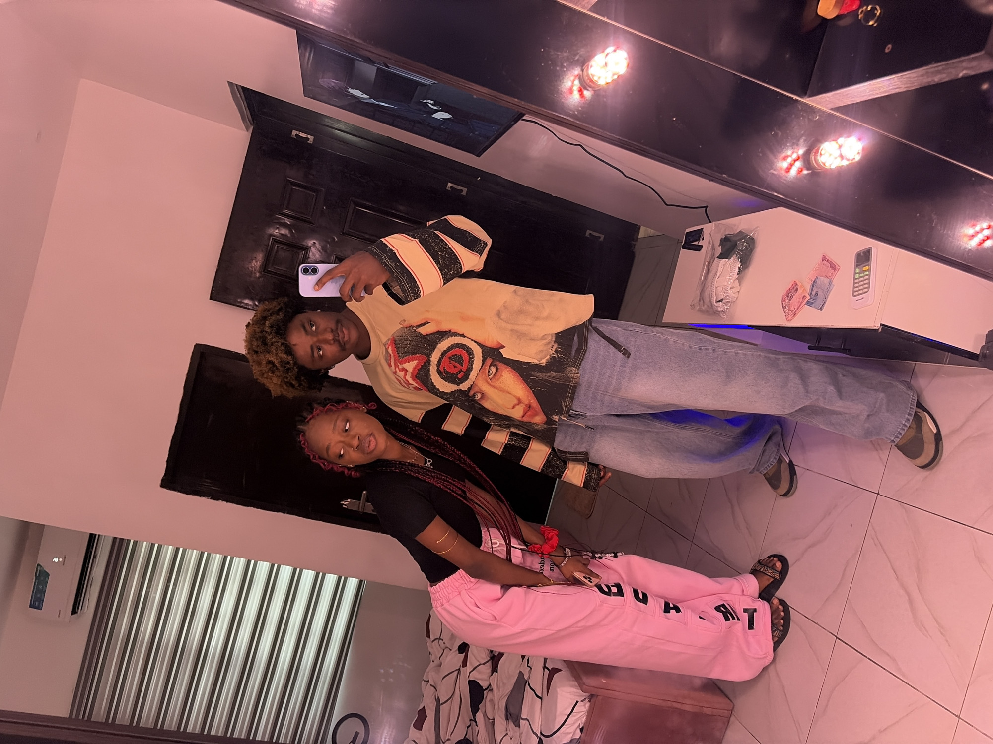 Two individuals are posing for a photograph in what appears to be an indoor setting, likely in Igboefon, Nigeria. One individual, possibly male, has curly brown hair and is wearing a graphic-printed beige sweatshirt and light blue jeans. He is holding a cell phone up, presumably to take the photo. The other individual, possibly female, has long braids and is wearing a black top and pink pants. They are positioned in front of a white wall and patterned curtains. In the background, there is a door. A vanity table with lights, what looks like a telephone, and money are visible. The floor is tiled.