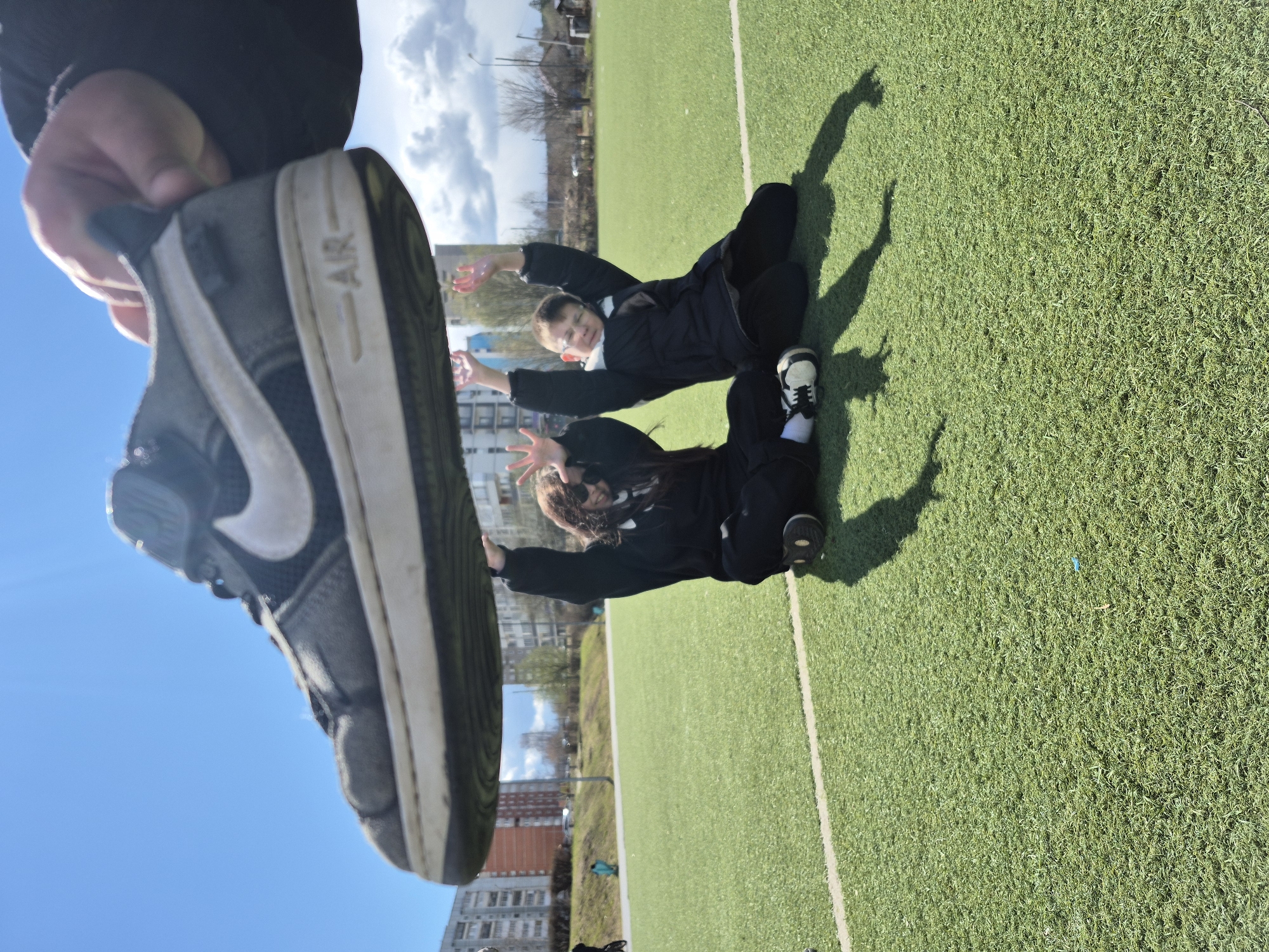 This outdoor photograph, taken in Chaikovsky, Russia, captures a low-angle perspective on an artificial turf sports field during daytime. The main subject in the immediate foreground is a person's hand holding a black and white Nike sneaker, with the word "AIR" distinctly visible on its white sole. The shoe is positioned very close to the camera, partially framing the scene behind it.
Further back on the bright green artificial turf, two individuals are seen lying down, appearing to pose playfully for the camera. Both are dressed in dark, likely black, attire and sneakers. One person, wearing glasses, is visible from the chest up, lying on their back with arms extended upwards. The second person, with long dark hair, is also lying down with arms raised, partially obscured by the foreground shoe.
The sunny weather is evident from the clear blue sky with scattered white clouds and the distinct, elongated shadows cast by people on the grass, suggesting a mid-day or afternoon setting. In the background, typical urban elements such as apartment buildings and some trees can be observed, indicating the location is likely a community sports facility. The image conveys a lighthearted, casual moment on a sunny day.