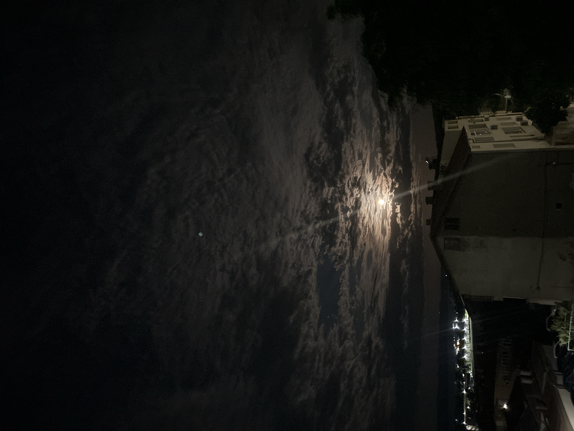 The image depicts a night scene with a cloudy sky dominating the view. A bright light source, likely the moon, is visible amidst the clouds. The scene includes buildings and urban structures. Distant lights suggest a cityscape, possibly along a coastline. The location appears to be in Burgas, Bulgaria. The image has been taken from an elevated position, with the view oriented vertically.
