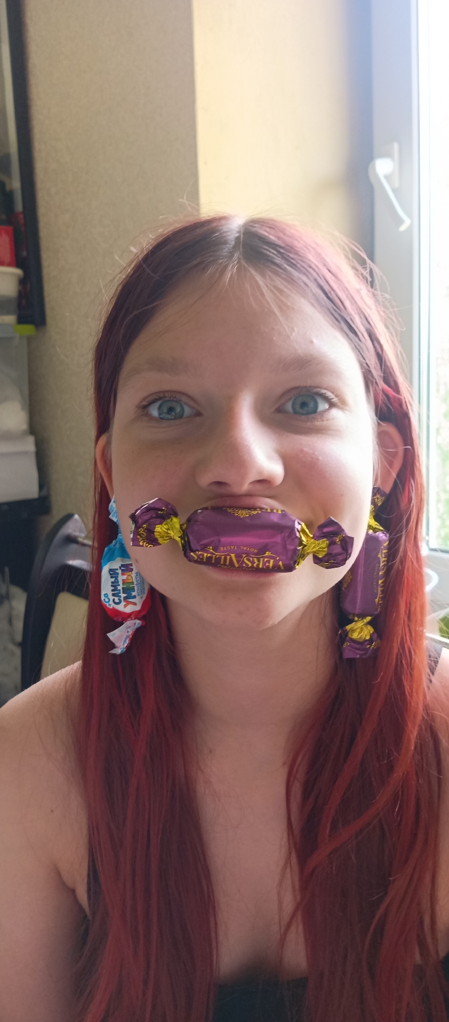 A young woman with light skin and blue eyes is centered in the frame. Her hair is long, red, and straight. She is holding a purple and gold "Versailles Royal Taste" wrapped candy in her mouth, horizontally between her lips. Dangling from her right ear is a similar candy. Dangling from her left ear is a "Co Самый Умный Конфеты" wrapped candy. Behind her, on the left side of the frame, is a partially visible cabinet with various items. To the right is a window. The location is Unknown, Unknown.