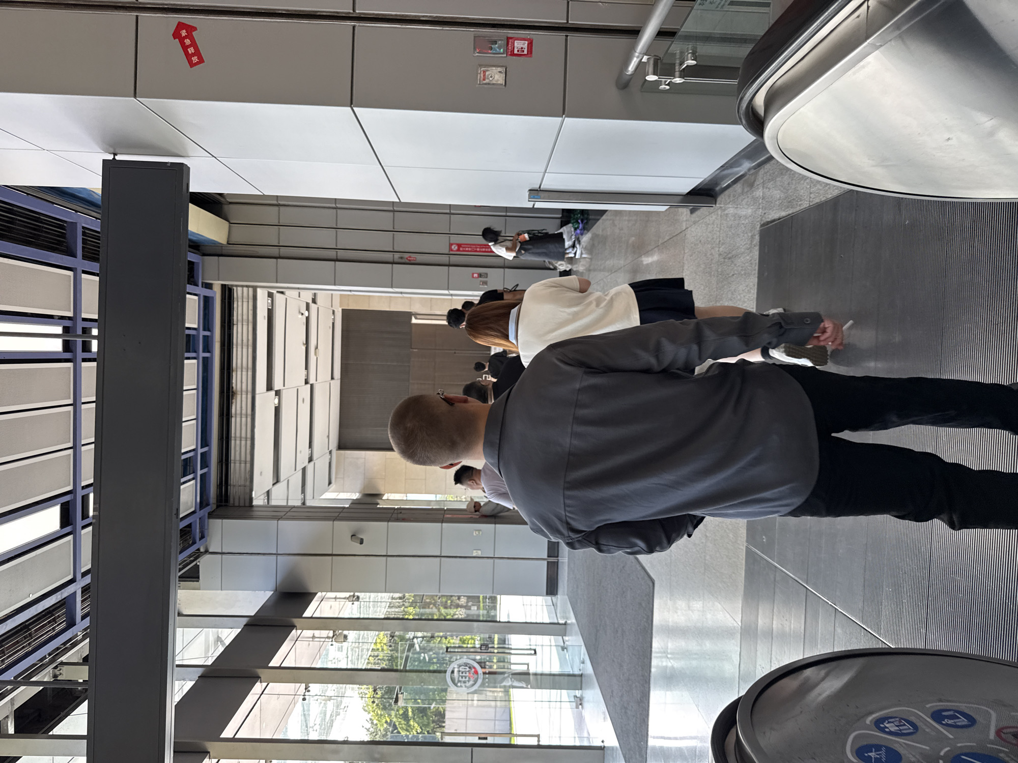 The image shows the interior of a modern building, likely a commercial or public space, in Haizhu District, China. In the foreground, two individuals are seen from behind, standing on a moving walkway or escalator. The person on the left is wearing a grey long-sleeved shirt and black pants, while the person to their right is wearing a white top and a dark pleated skirt. Behind them, a line of people is visible, heading towards an opening in the building's interior. The environment features polished floors, glass partitions, and architectural elements such as beams and ceiling panels. A red arrow sign with Chinese characters is visible on a pillar on the right side of the frame. In the distance, glass doors lead to an outdoor area with greenery. The overall impression is one of a clean, contemporary urban interior.