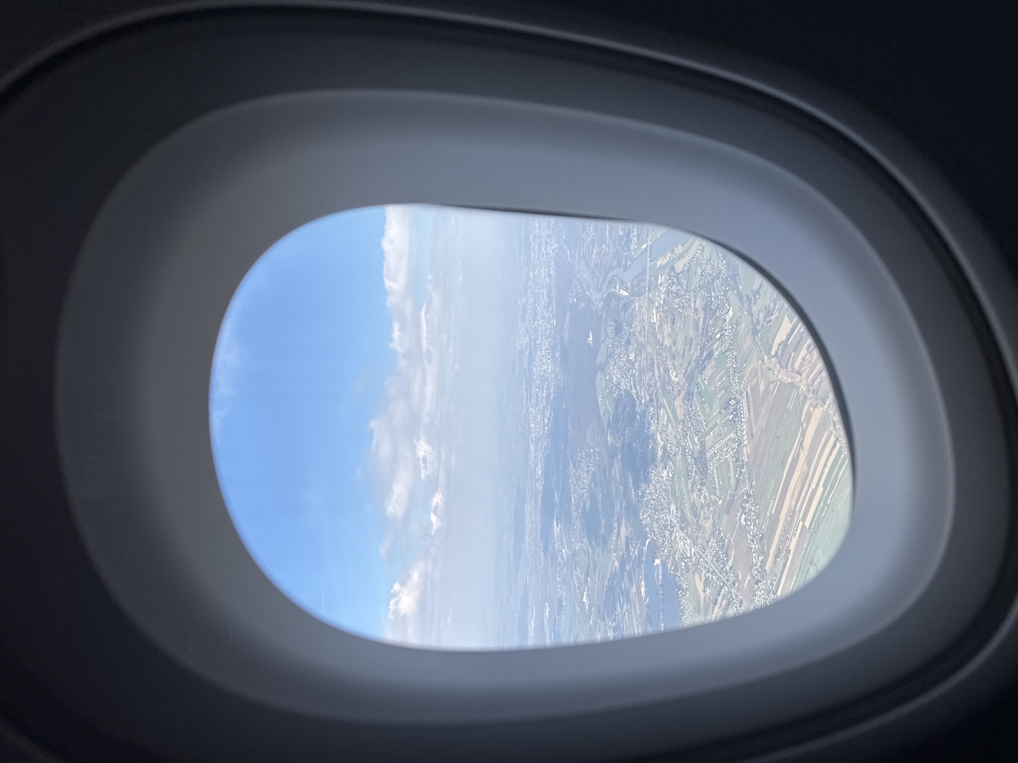 The image shows a view from an airplane window. Outside, there is a landscape featuring a mix of blue sky with white clouds and a view of land with fields, waterways, and sparse buildings. The land appears to be in a rural or semi-rural area, potentially near a city.
Given the prompt to identify the location in the city of Dąbrowa Szlachecka, Poland, it can be interpreted that the landscape visible from the plane window includes this city, or is in its vicinity.