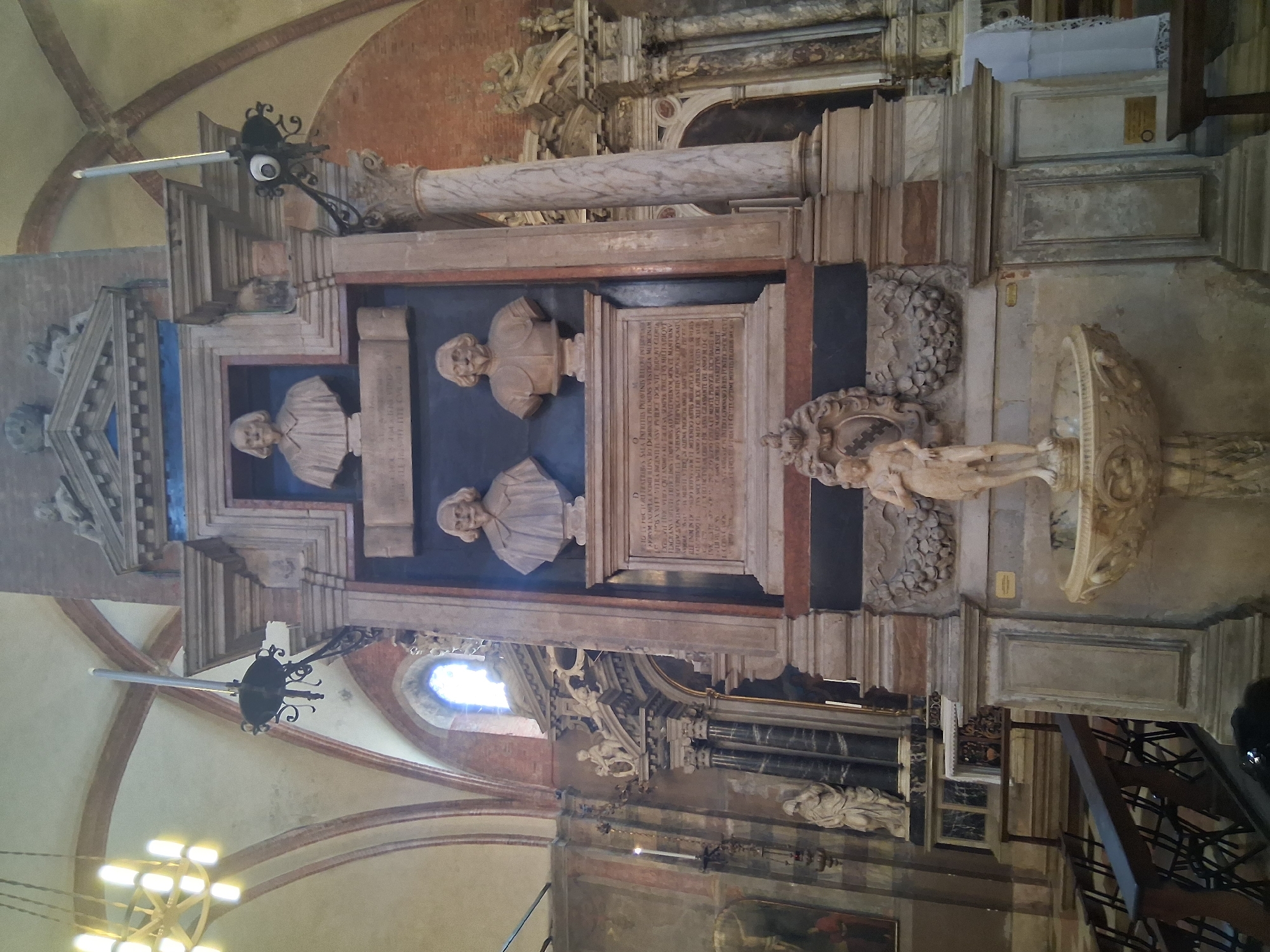 The image depicts a view inside a church, specifically the Basilica of Saint Anthony in Padua, Italy. The central focus is a large, ornate monument, likely a tomb or memorial, with several sculpted busts and intricate architectural detailing. The monument features at least three sculpted busts of men, one at the top and two below. A smaller, naked statue of a figure stands on a fountain at the base of the monument. The monument is flanked by large marble columns. In the background, architectural elements of the church are visible, including arched ceilings with exposed brickwork, stained glass windows, and other religious artworks and fixtures such as a chandelier and wall sconces with candles. The scene appears to be captured during daylight, with light streaming in from a window. There are no people clearly visible in the immediate foreground, but the presence of benches suggests this is an area accessible to visitors.
