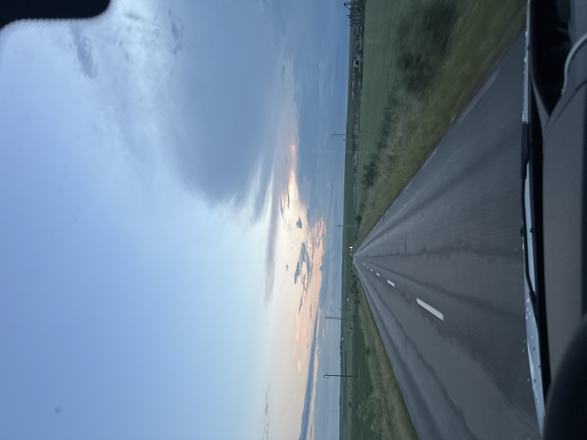 The image presents a view from a vehicle traveling along a straight, asphalt road, potentially near Zimnitsa, Bulgaria, though specific city elements are not visible. The road is bordered by grassy fields on either side, receding into the distance. Utility poles are spaced along the roadside. The sky is partly cloudy, with a mix of blue and areas of light orange and pink near the horizon, possibly indicating sunrise or sunset. The road's surface appears slightly wet, reflecting the ambient light. The passenger side of the vehicle is visible on the right.