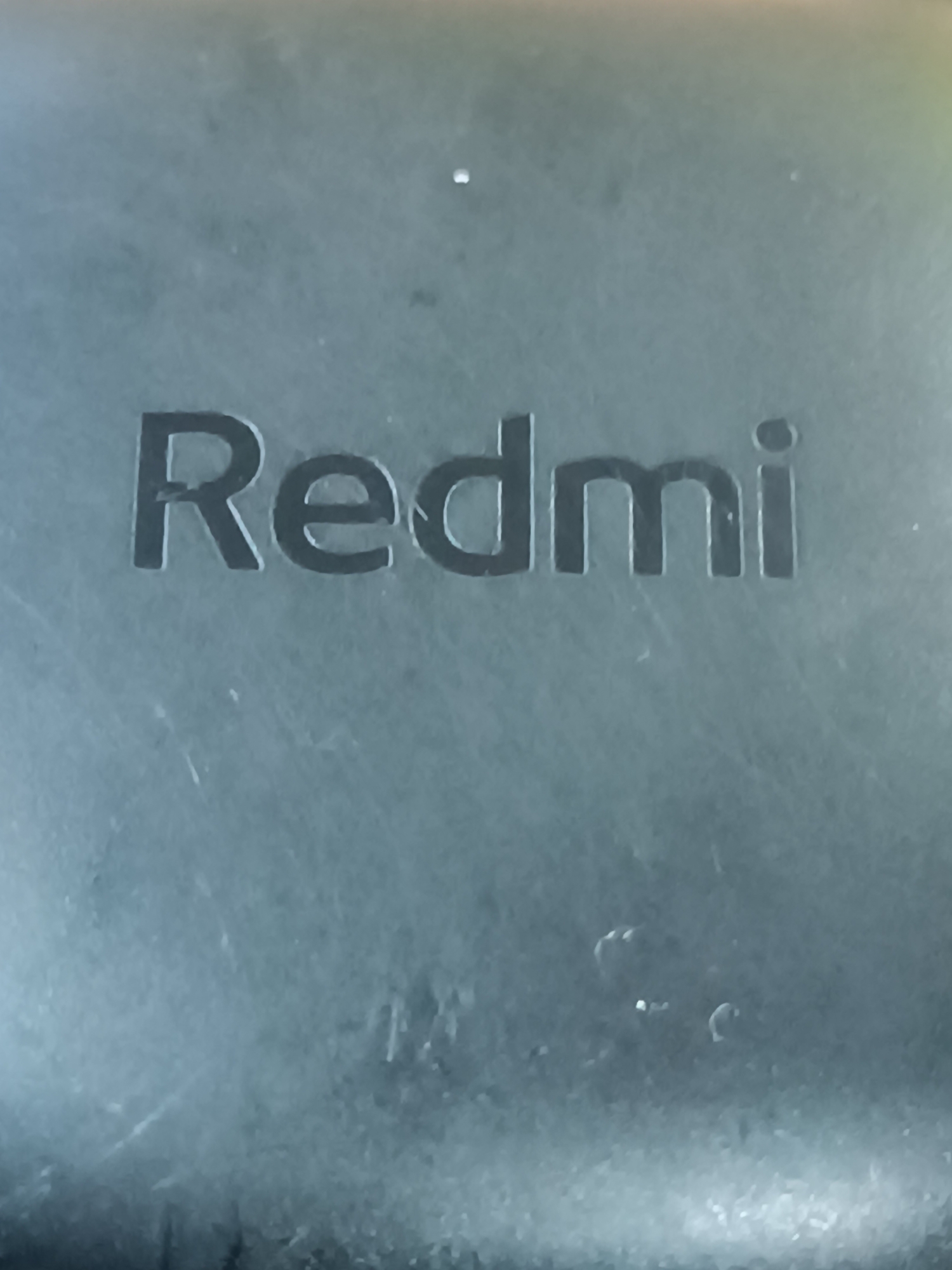 The image shows the word "Redmi" embossed on a dark, textured surface. The lettering is in a sans-serif font and appears to be a logo or brand name. There are some minor imperfections and scuff marks visible on the surface. There is no indication of the location being Khashuri, Georgia, or any other specific city. The image is a close-up of the text on an unspecified object.