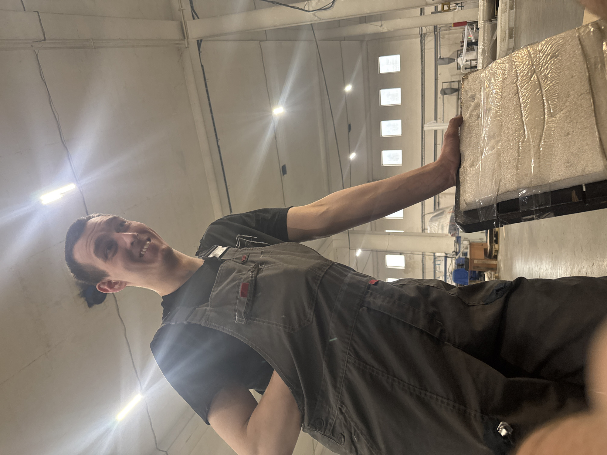 A male individual is depicted within a large industrial or warehouse facility in Dikaya Gar', Russia. He appears to be in his late twenties to early thirties, with dark hair styled in a small top bun and a faint goatee. He is smiling broadly, directly engaging the camera. He wears a black short-sleeved t-shirt beneath dark grey work overalls, which exhibit red accent tags on pockets and visible buttons on the straps. His left hand is casually inserted into a side pocket of his overalls. His right hand rests atop a rectangular object, approximately the size of a small panel, securely wrapped in clear plastic film. The material inside the wrapping appears to be light-colored and fibrous or granular, supported by a darker frame. The background features high, light-colored concrete walls and an exposed ceiling with multiple bright overhead fluorescent tube lights, creating significant lens flare. Distant square windows are visible high on a back wall, along with structural columns, and indistinct industrial equipment or stacked materials on a grey concrete floor.