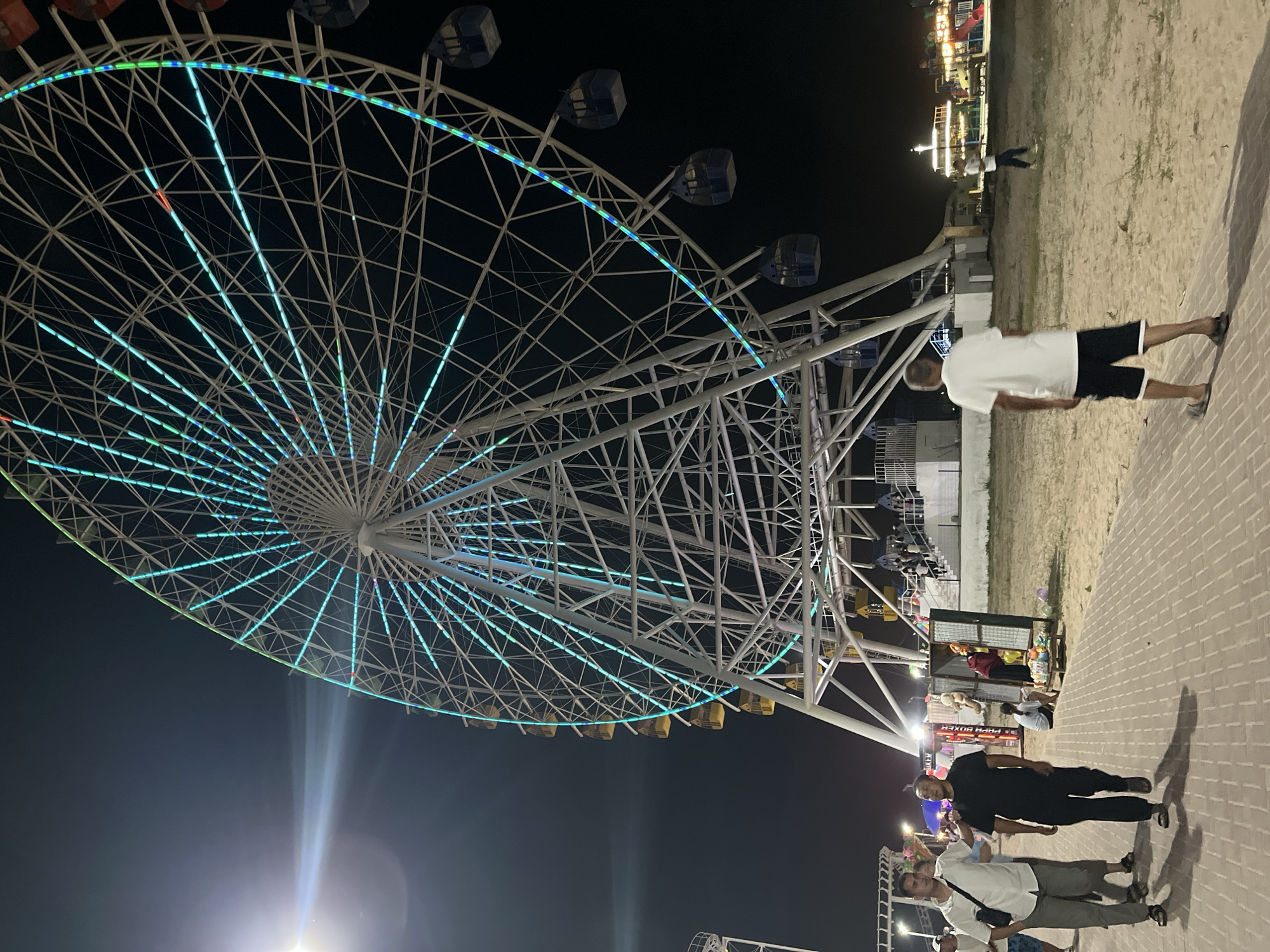 A large Ferris wheel illuminated with blue and green lights dominates the night sky in Bosteri, Kyrgyzstan. The wheel has multiple cabins, some of which are visible as dark blue structures. In the foreground, a paved walkway runs through a sandy area. Several people are walking on the walkway and in the sandy area. On the left, two men are walking towards the viewer, one in a white shirt and light-colored pants, the other in a black t-shirt and black pants. Further back, a man in a white t-shirt and black shorts is walking away from the viewer. In the background, there are food stalls, other amusement rides, and more people. The overall scene depicts a lively amusement park at night.