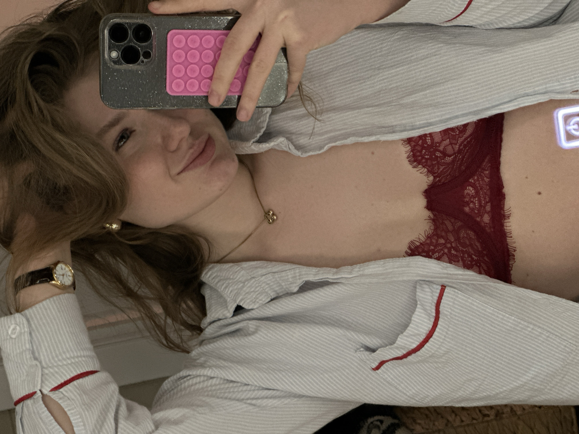 A woman is taking a selfie. She is wearing a light blue and white striped pajama top with red trim, and a red lace bra. She is holding a smartphone with a pink pop socket. A gold watch is visible on her left wrist, and a gold necklace is around her neck. The background is blurred and indistinct, making it impossible to identify the location. The image does not provide any evidence of it being in Kazan, Russia.