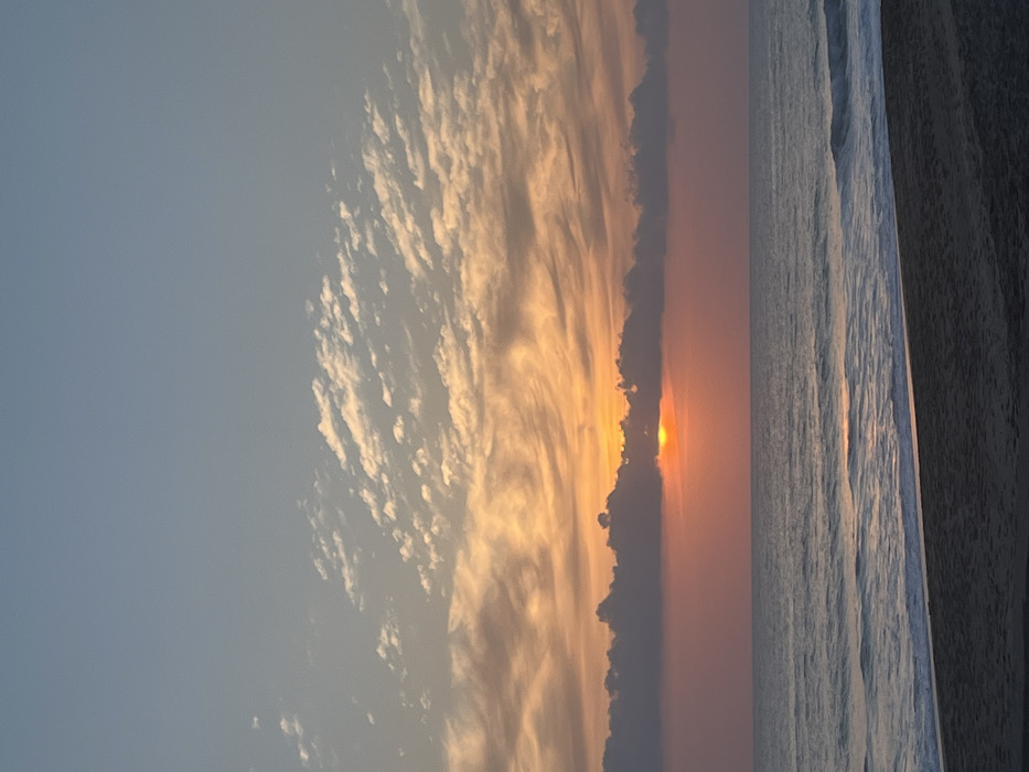 The image displays a coastal scene in Alanya, Turkey, capturing either sunrise or sunset over the Mediterranean Sea. The horizon is flat and clearly defined. The sun, a bright orange-yellow disc, is positioned low and centrally, partially obscured by a horizontal band of dark grey clouds directly above the water. Above these clouds, the sky exhibits a gradient, transitioning from light blue at the top through layers of scattered, streaky white clouds, into a broader band of orange and pink hues that glow around the obscured sun. The sea surface is calm, reflecting the sky's colors with muted blues and greys, and shows a distinct, shimmering path of sunlight extending towards the viewer. Gentle waves break with white foam at the water's edge, meeting a strip of dark, wet sand in the immediate foreground. No individuals, structures, or distinct objects are present within the frame.