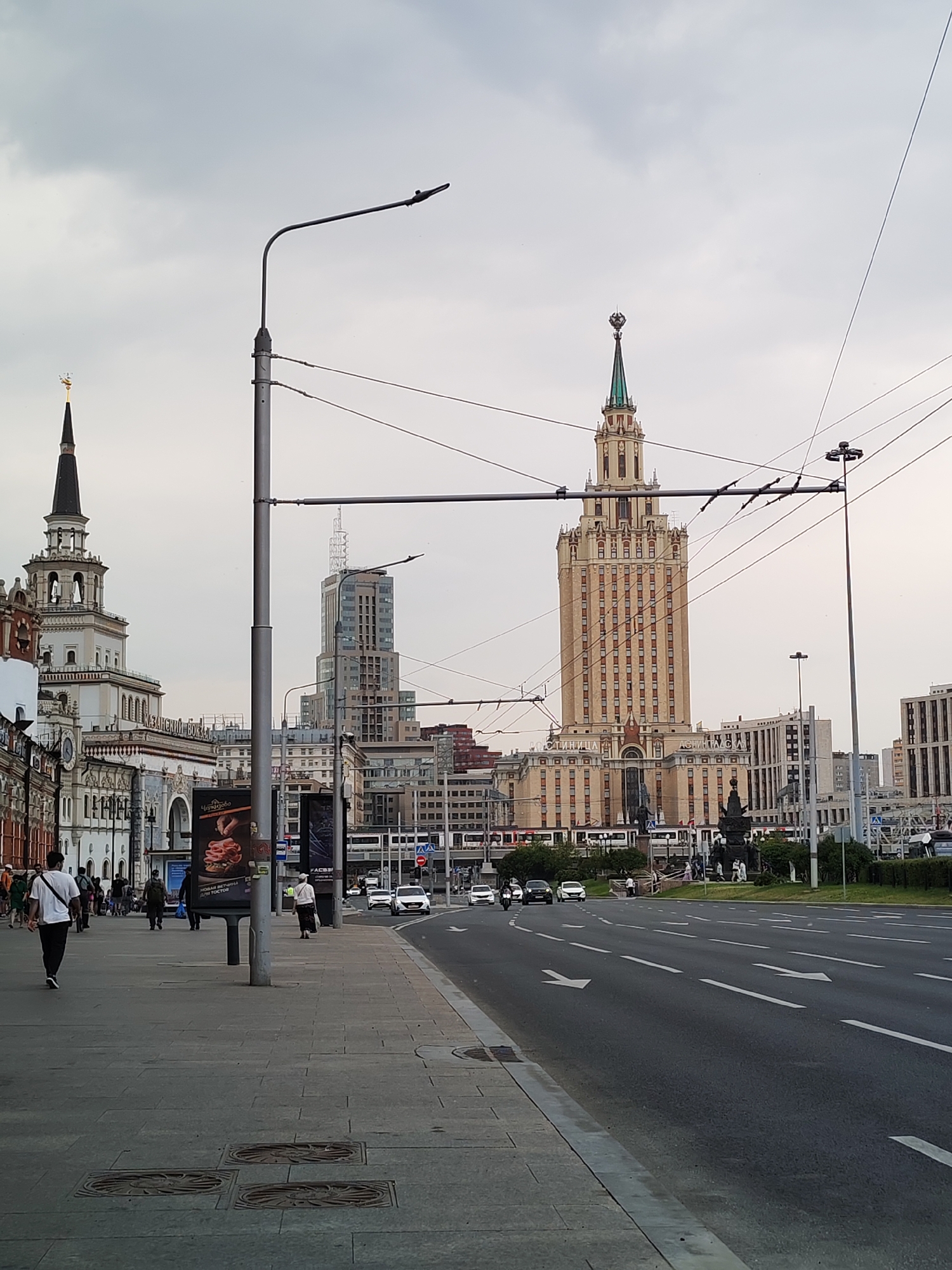The image depicts a street scene in Moscow, Russia, likely near Komsomolskaya Square. In the foreground, a paved sidewalk runs alongside a multi-lane road. Several people are walking on the sidewalk, some appearing to be tourists. Behind them, a collection of buildings is visible, including the iconic Stalinist skyscraper, Hotel Ukraina, and the historic Kazansky railway station. The sky is overcast, suggesting a cloudy day. Vehicles are moving on the road, and overhead power lines are visible, indicating the presence of public transportation like trams. Advertisements are displayed on poles along the sidewalk. The overall atmosphere is that of a busy urban environment.