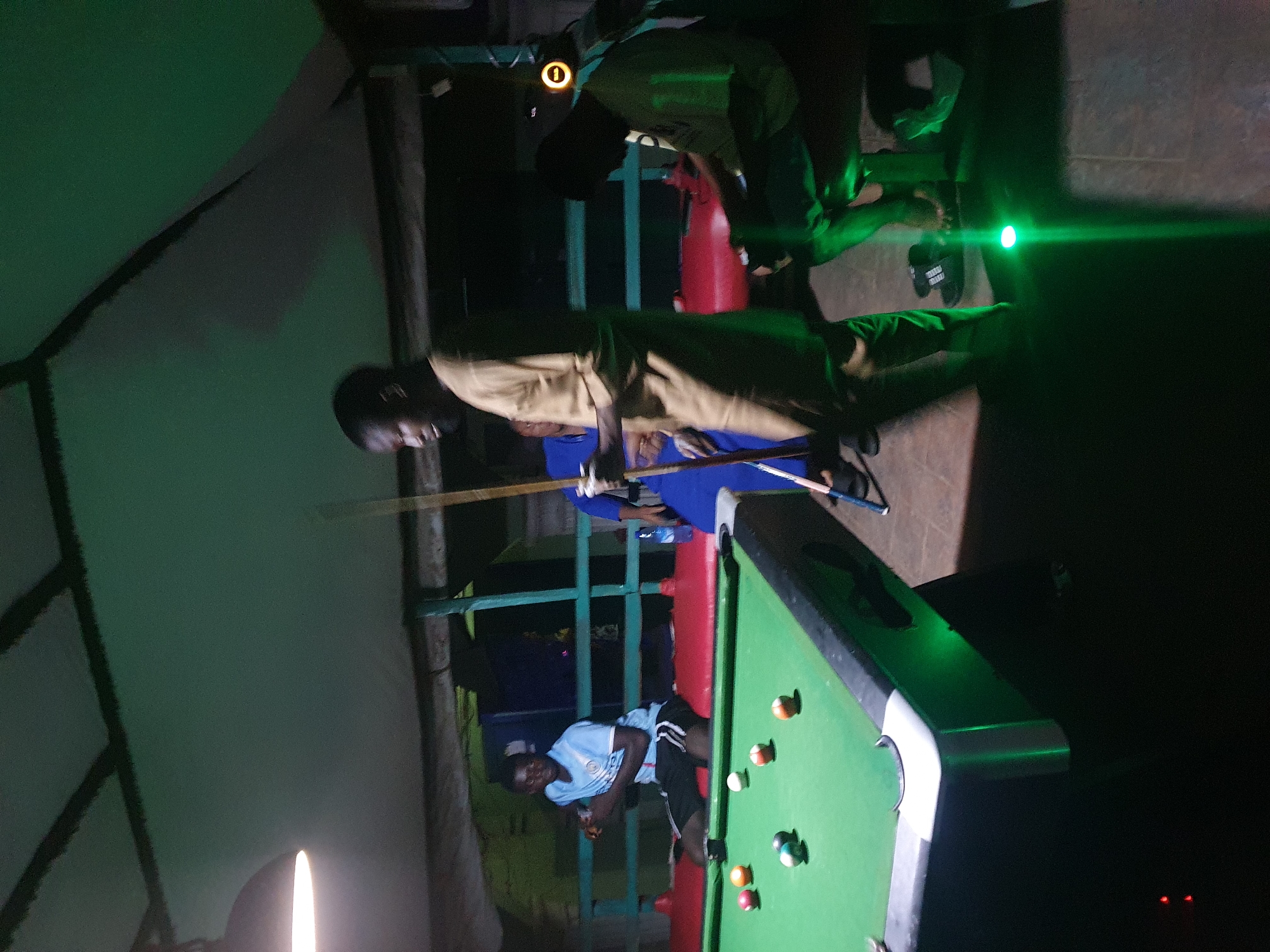 The image depicts an indoor pool hall in Lokoja, Nigeria, at night. The scene is dimly lit, with green and white lighting casting an atmospheric glow. In the foreground, a pool table with green felt and multi-colored balls is visible. Several men are present, engaged in a game of pool. One man, wearing a light blue shirt and black shorts, is positioned on the table, ready to play. Another man, in a tan outfit, stands to the right, holding a pool cue. A third man, in a green shirt, is seated on the left side of the table. The environment suggests a recreational setting, possibly a bar or club, with a suspended ceiling and exposed beams. The overall impression is one of a casual gathering.
