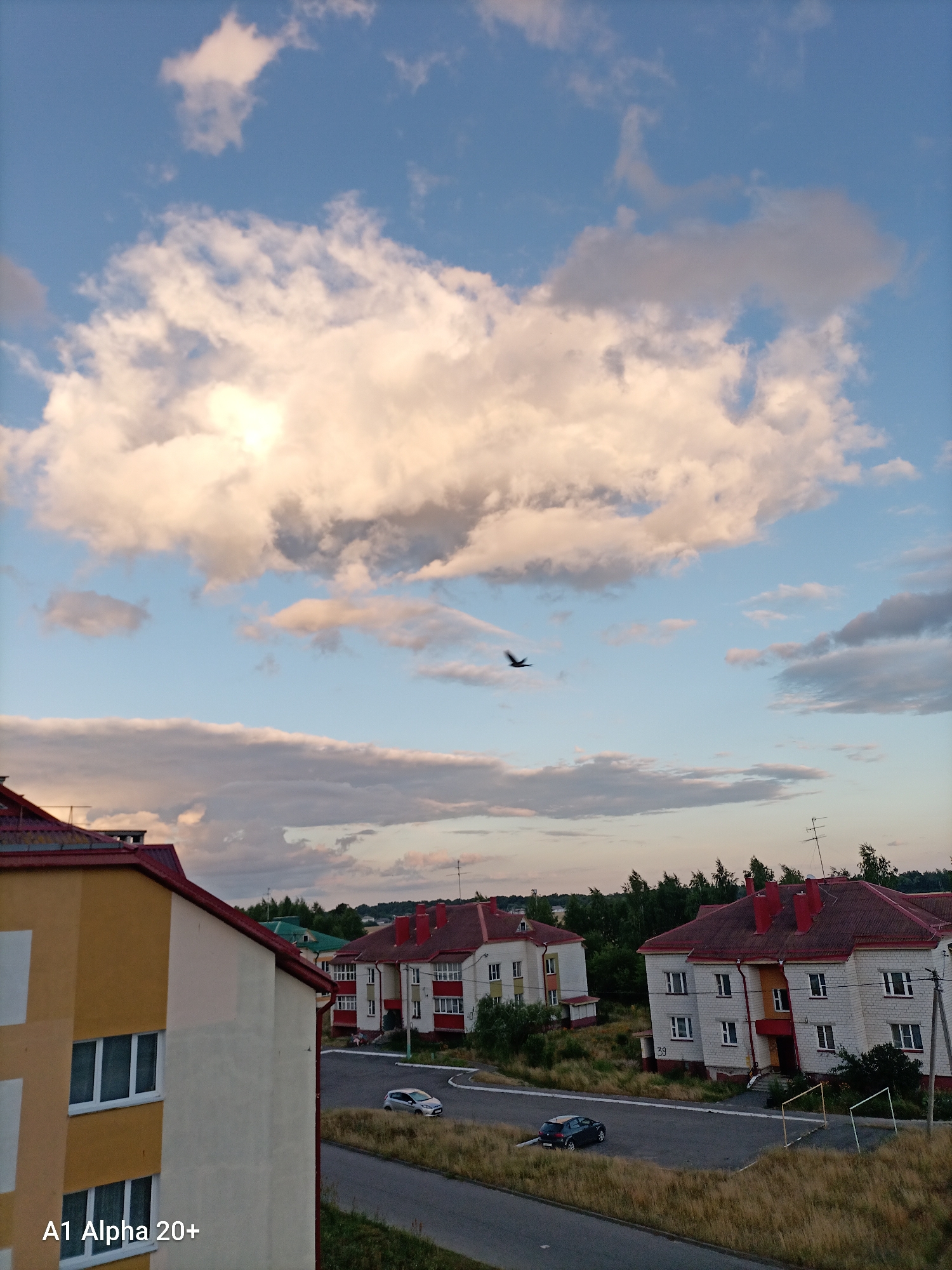 The image captures a scene in a residential area, likely in the Улукаўскі сельскі Савет, Belarus, during what appears to be late afternoon or early evening. A large, fluffy white cloud dominates the upper portion of the sky, with patches of blue visible around it. A dark bird is in flight in the middle ground of the sky. Below the sky, several multi-story apartment buildings with red roofs are visible. The buildings are constructed with light-colored brick and feature numerous windows. In front of the buildings, a paved road or driveway curves through a grassy area. Two cars are parked on the road. A tall, thin antenna is mounted on one of the buildings. The foreground features a portion of a yellow and cream-colored building with visible windows, and the text "A1 Alpha 20+" is superimposed in the lower left corner. The overall impression is of a typical urban or suburban residential setting under a partly cloudy sky.