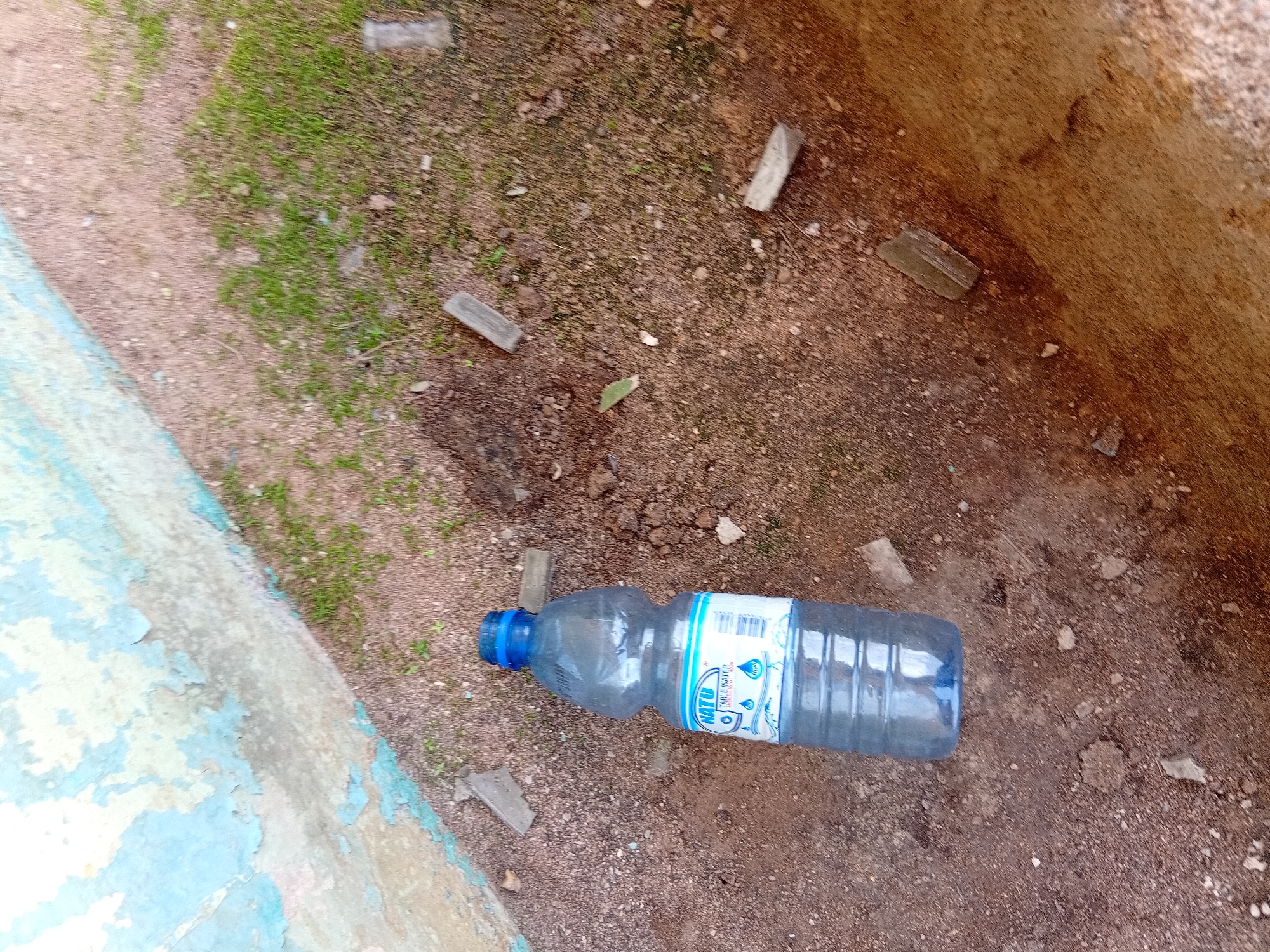 A partially full plastic bottle of "NATU TABLE WATER" rests on the ground, surrounded by small pieces of broken glass. The ground appears to be a mix of dirt, small stones, and sparse green grass. To the left, a wall with peeling blue and white paint is visible, suggesting an outdoor urban or semi-urban environment. The scene is likely located in Jalingo, Nigeria, given the context provided. There are no people or animals present. The lighting suggests it is daytime.