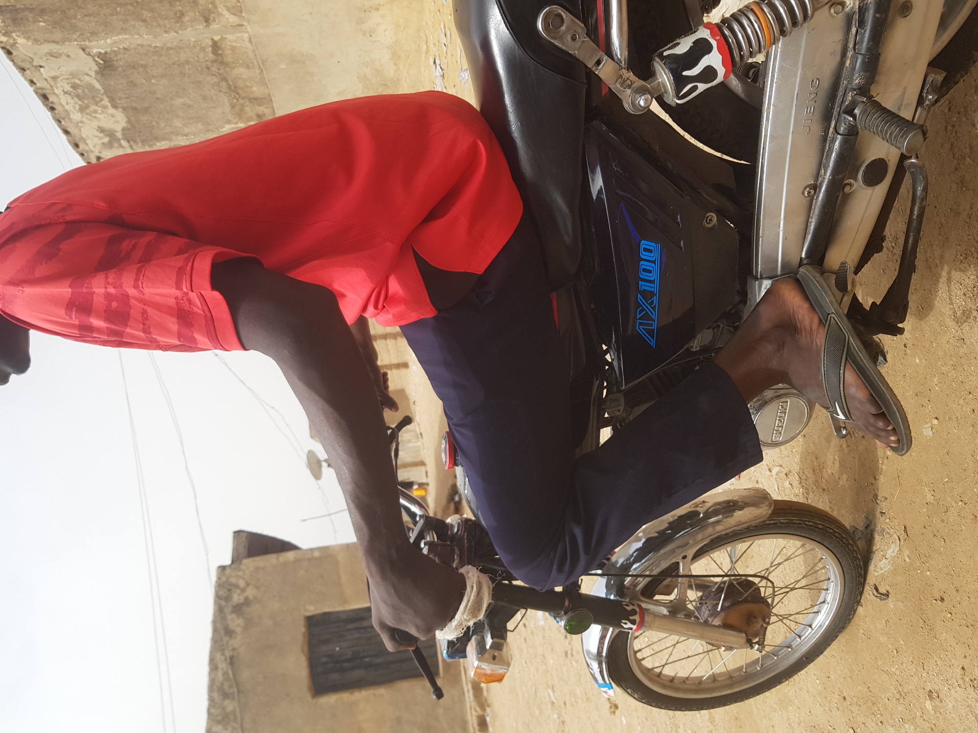 An individual wearing a red shirt and dark blue pants is seated on a Suzuki motorcycle, model AX100. The person is wearing a grey flip-flop on their right foot. The left hand is gripping the handlebar, which is wrapped with a white bandage. The setting appears to be Dutsinma B, Nigeria, suggested by the surrounding architecture. A building with a window is visible in the background. The ground appears to be dirt.