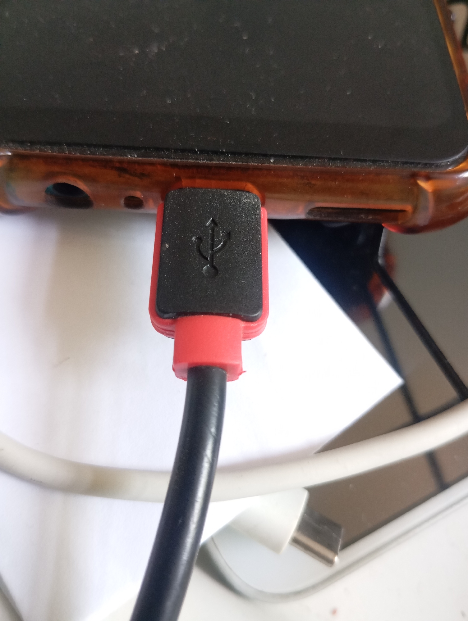 The image shows a close-up view of a USB cable plugged into the port of a smartphone. The phone has a dark screen and is housed in a translucent orange case. The USB connector is black with a red rubber accent, and it features the universal USB symbol embossed on its surface. A black cable extends from the connector. In the background, a white cable and part of another electronic device are visible. The lighting suggests an indoor setting. The image focuses on the connection point of the charging cable and the phone, implying the activity of charging the device. There is no discernible text in the image.