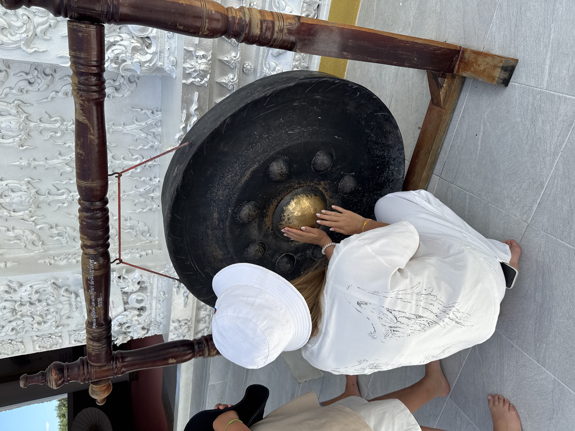 A person in white clothing and a white bucket hat is kneeling and touching a large gong. The gong is suspended from a wooden frame. The background features intricate white stucco decorations. The scene appears to be outdoors, possibly in a temple or cultural site. The person's hands are positioned on the central golden portion of the gong. The ground is tiled.