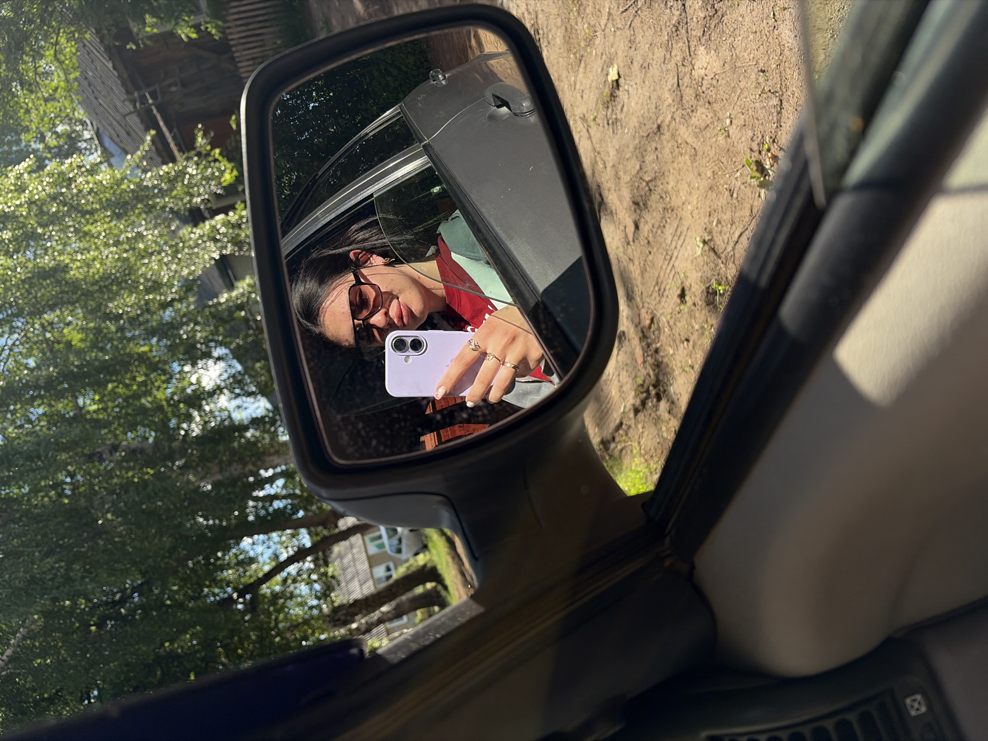 A woman is reflected in a car's side mirror. She is wearing sunglasses, a red jacket over a green shirt, and holding a purple cell phone in her right hand. Trees and a building are visible in the background outside of the mirror. The ground outside the car is dirt. The location might be Фоки, Russia based on the information provided.