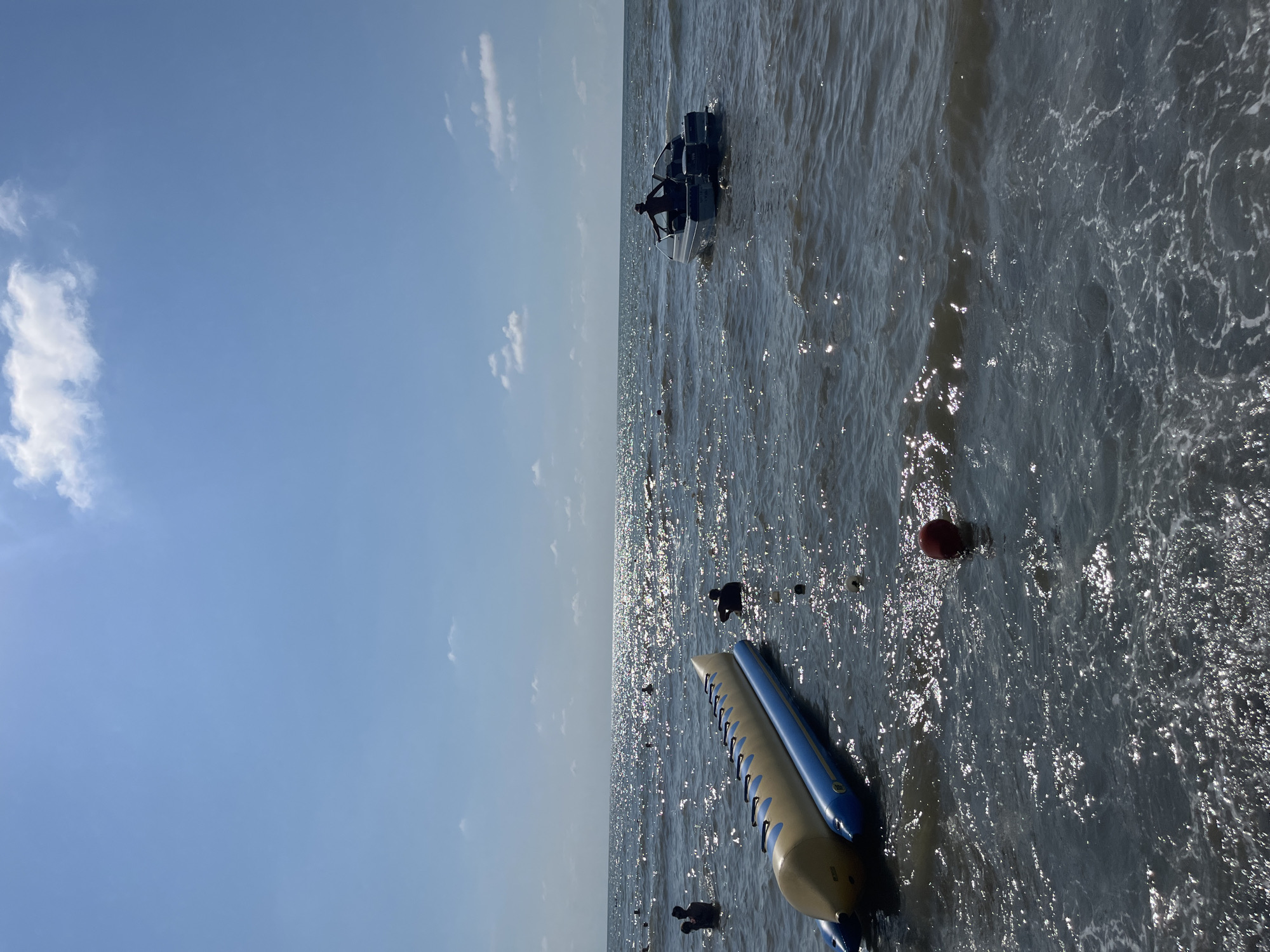The image depicts a sunny day at the beach in Ureki, Georgia. The sea is calm with gentle waves, and the sun sparkles on the water's surface. In the foreground, a yellow and blue inflatable banana boat is partially submerged. To the right, a small motorboat with a person standing in it is cruising on the water. Several people are visible in the sea, some swimming and others wading. The sky is clear blue with a few scattered white clouds. The scene suggests a leisurely day at a popular seaside resort.