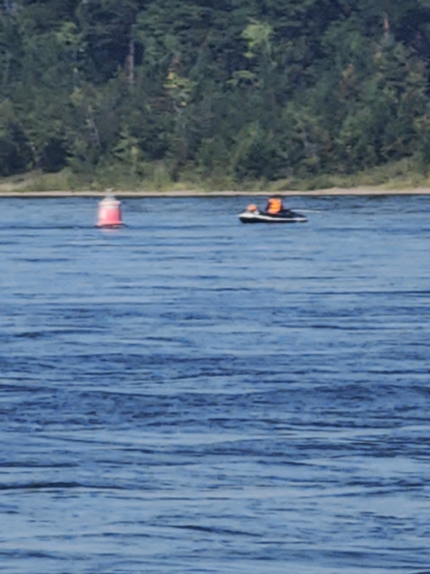 In the foreground, the deep blue water of a wide river is textured with ripples. In the middle ground, a bright pink and white buoy floats on the surface. Further back, a black and white jet ski navigates the water. Two individuals, wearing bright orange life vests, are seated on the jet ski. In the background, a dense, green forest lines the distant shore. The overall scene is one of recreational activity on a body of water, with a natural landscape as the backdrop. The location is the city of Ермолаево, Russia.