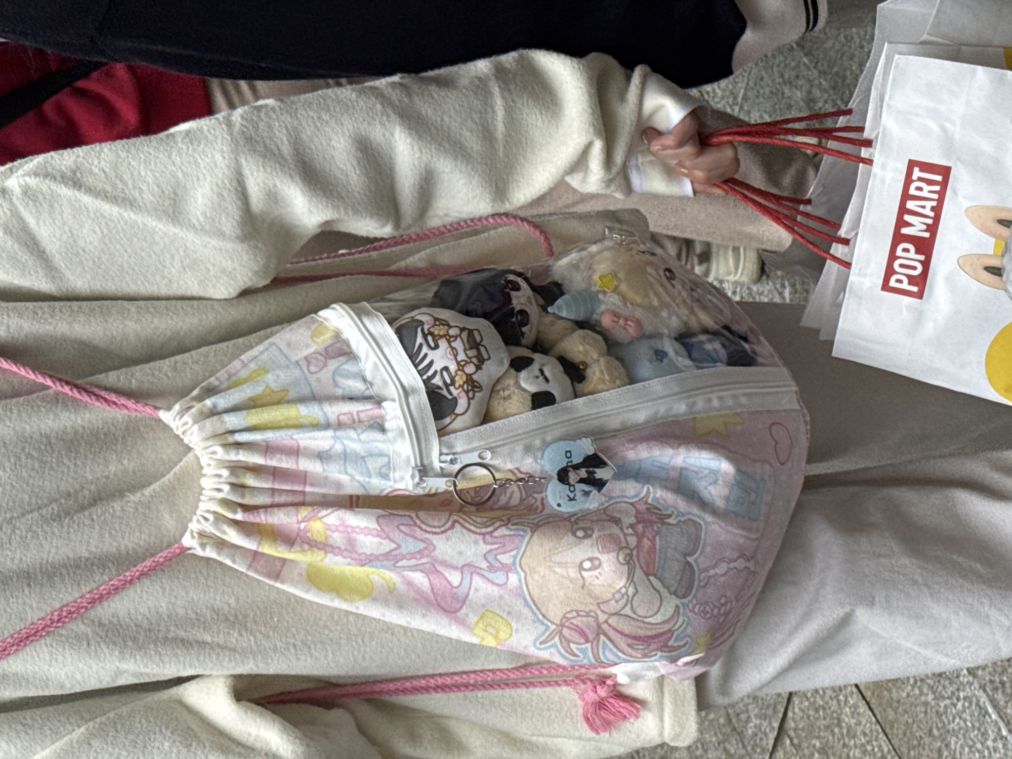 The image shows a person holding shopping bags from "POP MART," a retail store known for selling collectible toys and art toys. The bags have red handles. Nearby is a drawstring bag with a zipper and featuring cartoon character prints in pastel colors. The bag is filled with plush toys. The backdrop consists of a light-colored fabric or blanket.
There is no information about the location being in Jinjiang District, China.