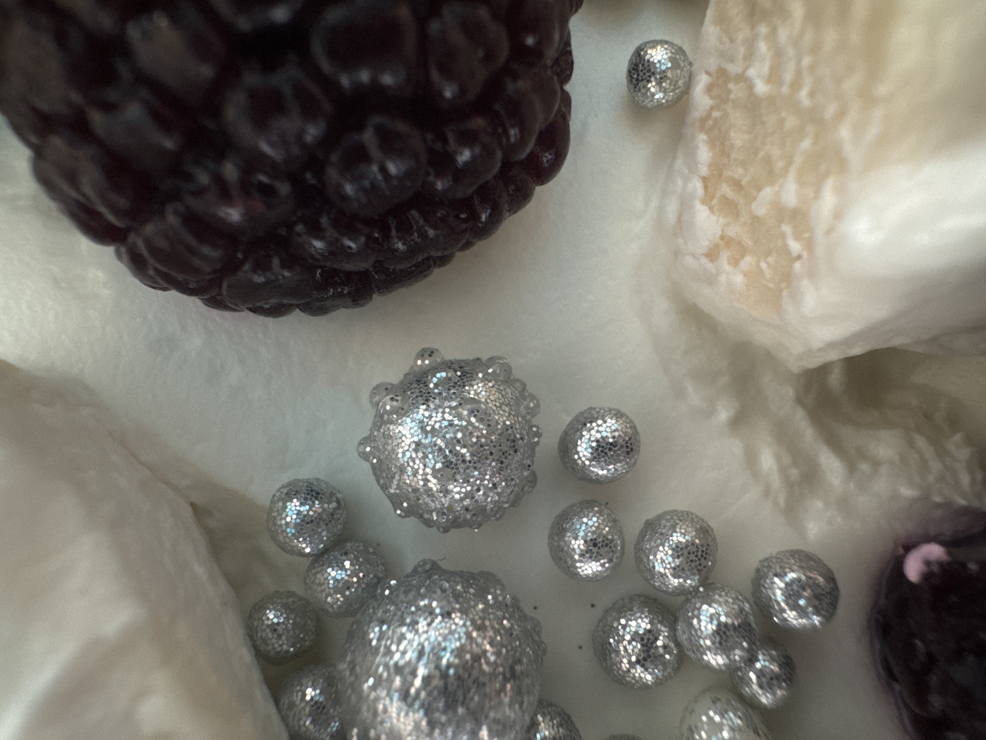 This image, originating from the city of Chaikovsky, Russia, presents a high-angle, close-up still life composition. Approximately two dozen silver, glitter-covered spherical objects, ranging in size from roughly 1 cm to 4 cm in diameter, are scattered across the left and central areas of the frame. These spheres display varied textures; some are smooth, while others possess a distinctly bumpy or nodular surface. A prominent, large, dark-colored object resembling a blackberry or mulberry, characterized by its textured, multi-lobed structure, occupies the upper right quadrant. The scene is further delineated by two significant, irregular masses of light-colored, soft material: one is positioned in the upper left, and another, with a visible beige hue and textural variations, is in the lower right. The overall background is a continuous, light-toned, soft surface.