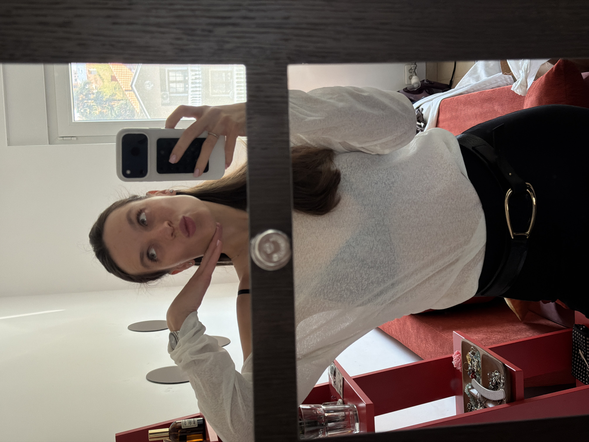 A young woman takes a selfie in a mirror. She is wearing a white, long-sleeved shirt and black pants with a gold buckle belt. A window is visible in the reflection, showing a view of buildings. A red storage unit with open shelves sits below the mirror, holding various items. The location is within Vozdovac Urban Municipality, Serbia, as evidenced by the architectural style visible through the window.