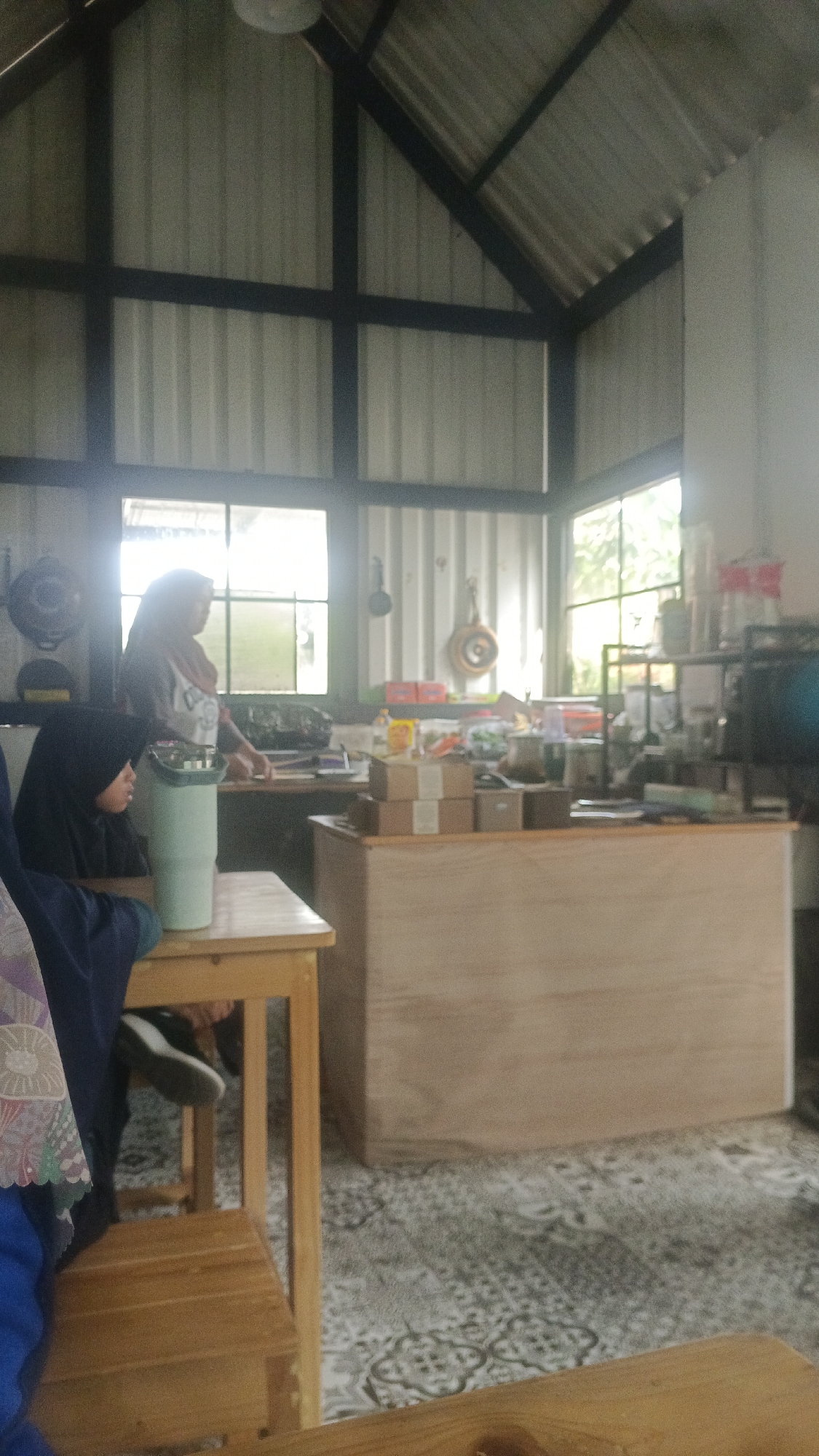 The image shows an indoor scene with two women. One woman, wearing a hijab, is seated at a wooden table. The other woman, also wearing a hijab, stands behind a counter, appearing to be working. The environment suggests a cafe or shop. The room has a high ceiling with exposed metal beams and a corrugated metal roof. Large windows allow natural light to enter. The counter area is stocked with various items, including boxes, containers, and what appear to be food or beverage products. The floor is covered with a patterned carpet. The scene is set in a casual, possibly commercial, space within Tangerang, Indonesia.
