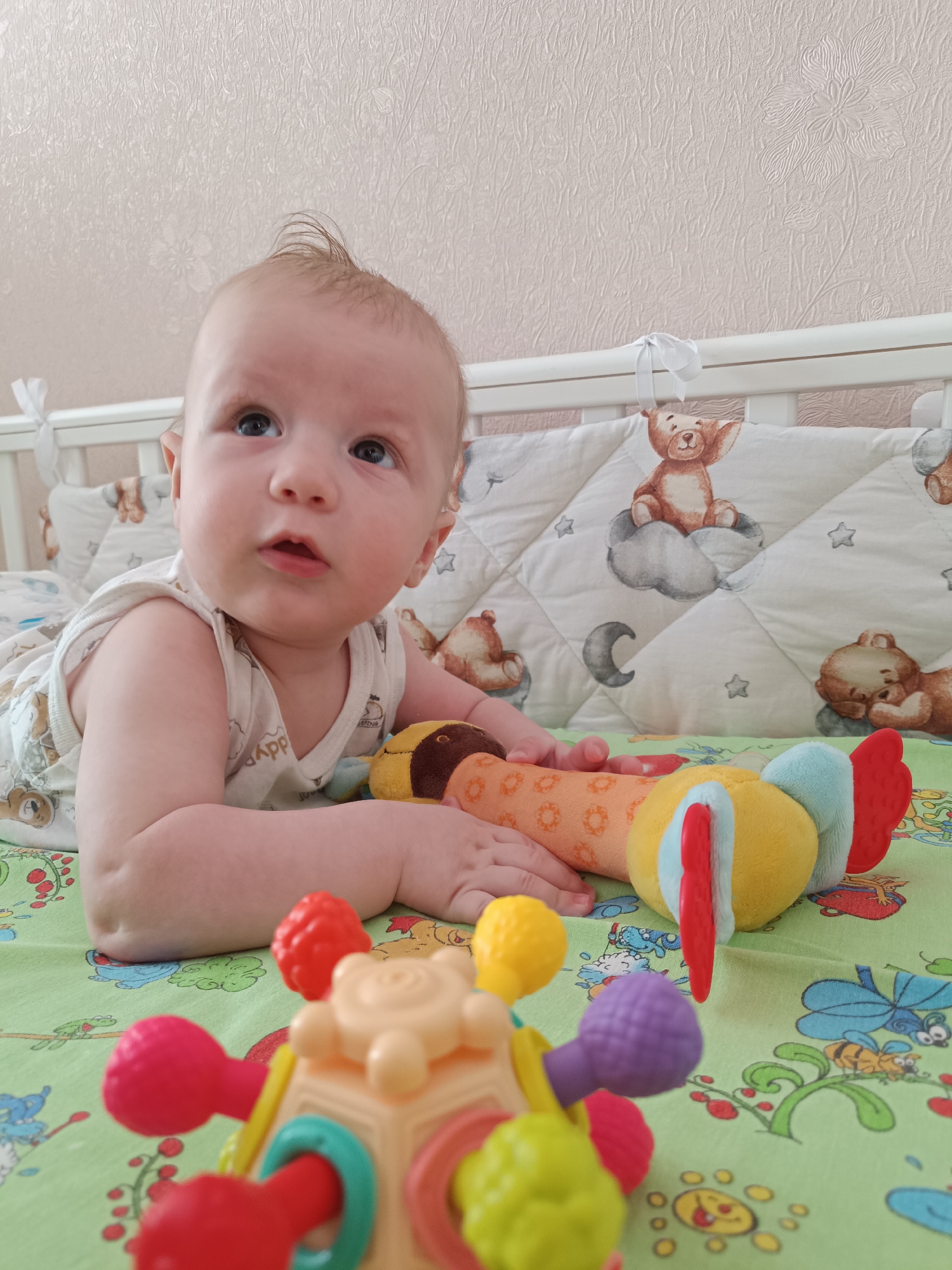 A baby is lying on its stomach in a crib. The crib has a green sheet with cartoon illustrations. There's a padded bumper around the inside of the crib, featuring a design of teddy bears on clouds and stars. The baby is wearing a white tank top with small light-colored pictures and appears to be looking upwards.
The baby is grasping a yellow and orange stuffed toy shaped like an animal. In the foreground is a colorful plastic toy with various protrusions.
The location is potentially a home in the city of Tobolsk, Russia. This conclusion is based on information provided by the prompt but is not directly discernible from the image itself. The background features a plain, light-colored wall.