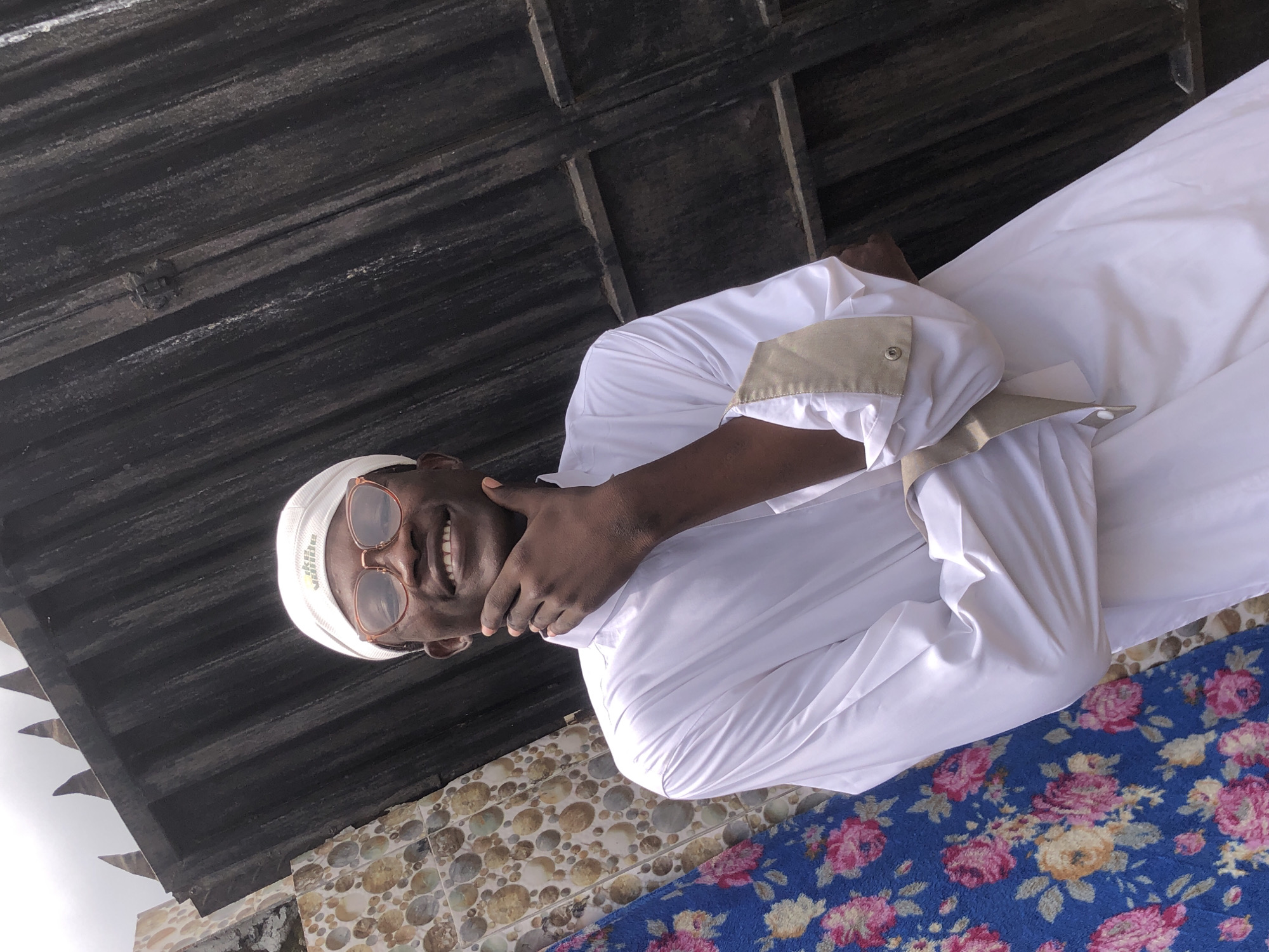 A young Black man wearing a white Kufi cap and eyeglasses smiles, resting his chin on his left hand. He is wearing a white thobe, which is a long, loose-fitting robe. The man is standing in front of a dark, corrugated metal fence. To the left of the man is a section of tiled wall and a blue fabric with a floral pattern. The overall setting appears to be outdoors, possibly in Ado, Nigeria.