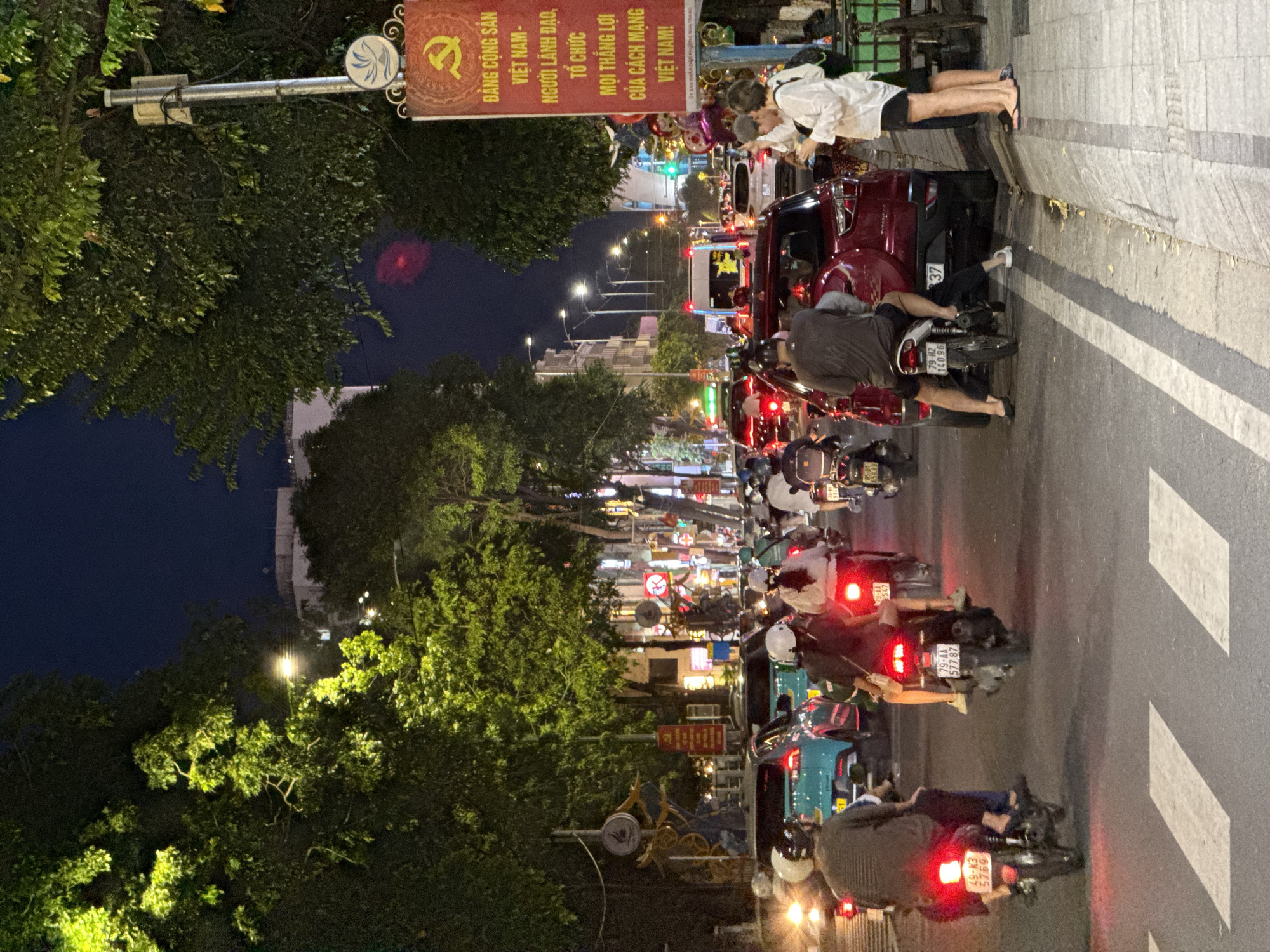 The image depicts a busy city street scene at night in Nha Trang, Vietnam. The street is filled with traffic, primarily motorcycles, with a few cars visible. Many people are riding on motorcycles, some with helmets and others without. The street is lined with trees illuminated by streetlights, creating a vibrant and somewhat chaotic atmosphere. In the background, buildings and signs can be seen, some with illuminated lettering. A prominent red banner with Vietnamese text and the symbol of the Communist Party of Vietnam is displayed on the right side of the image. The scene suggests a lively urban environment with active nightlife and transportation.
