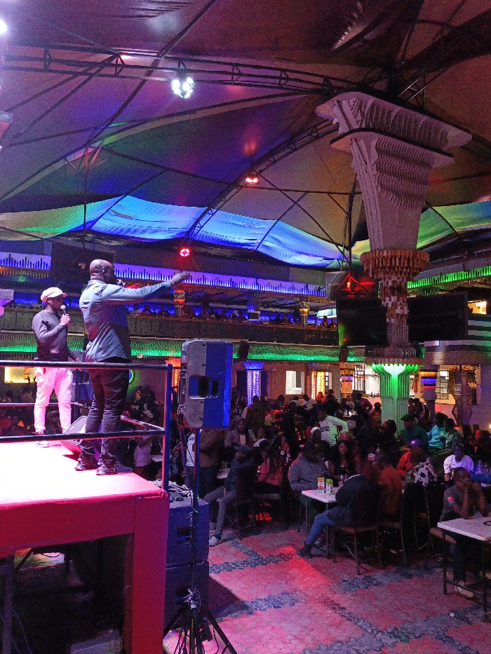 A vibrant nightlife scene unfolds with a stage hosting two performers, one in white pants and a hat, the other in a blue shirt pointing to the audience. A crowd fills the space, seated at tables under colorful lights and unique structural pillars. A balcony overlooks the lively scene. The floor has a patterned design. A speaker stands near the stage.
