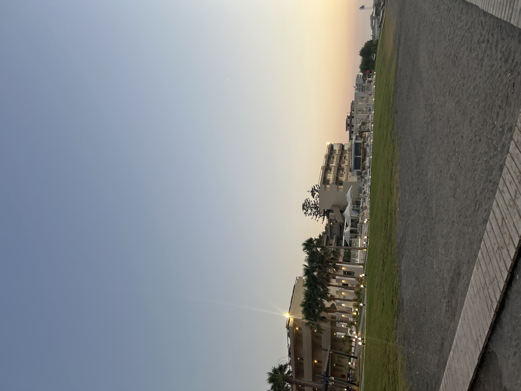 The image depicts a seaside resort area in Rhodes, Greece, likely during the late afternoon or early evening as indicated by the soft, warm light of the setting sun. The sky transitions from a clear, pale blue on the left to a gradient of orange and yellow towards the horizon on the right.
The scene is dominated by a long, paved walkway bordering a strip of green grass. Beyond the grass, a multi-story resort building is visible, characterized by white or light-colored facades, numerous balconies, and rows of windows. Palm trees are interspersed along the front of the resort, adding to the tropical ambiance. String lights are visible along the resort's exterior, suggesting it is preparing for evening illumination.
In the background, other resort buildings and some trees can be seen. A flagpole with a Greek flag is also present, reinforcing the location. The ground in the foreground appears to be a mixture of sand and gravel, typical of a beach environment. There are no people visible in the frame. The overall mood is peaceful and serene, capturing the tranquil atmosphere of a coastal destination at dusk.