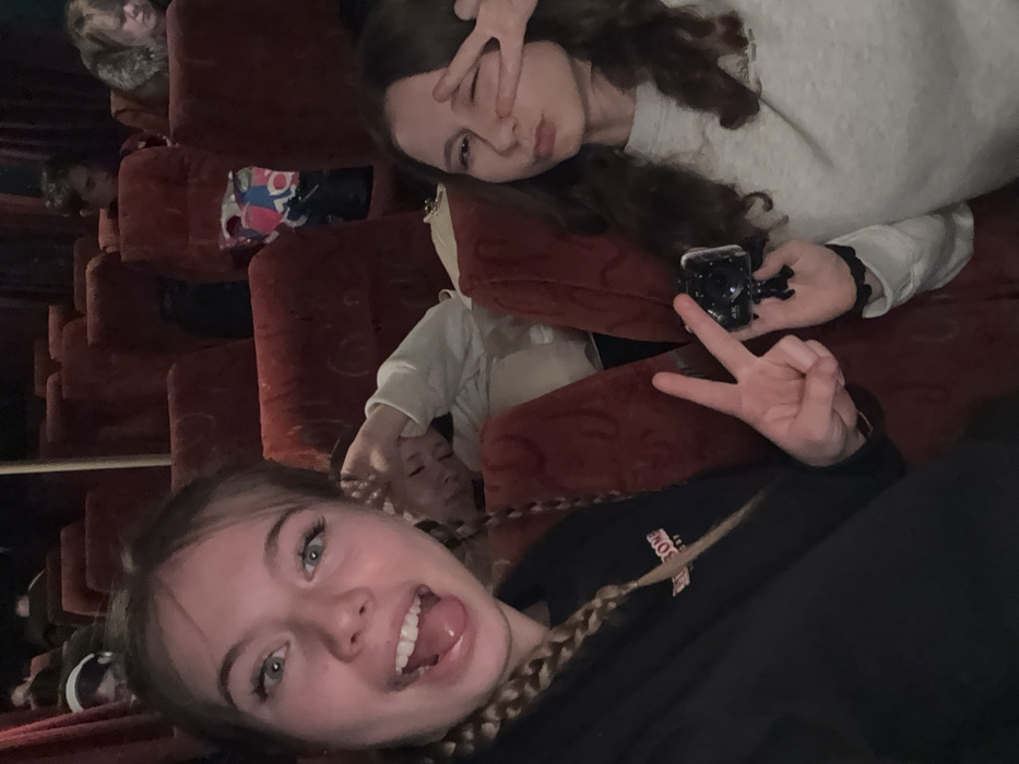 An image taken inside a passenger vehicle, likely a bus or coach, in Chernovskoe, Russia, shows multiple individuals seated in rows of red, upholstered seats under dim lighting conditions.
In the foreground, two young females are posing for the camera. The female on the left has light-colored hair styled in braids, prominent blue eyes, and is wearing a dark top. She displays an open-mouthed expression with her tongue slightly extended and makes a "peace sign" gesture with her left hand. The female on the right has dark, wavy hair and is wearing a light-colored long-sleeved top. She is captured with pursed lips and her right eye winking, while her right hand forms a "peace sign" over her winking eye. She holds a small, black action camera, possibly a GoPro, in her left hand.
Immediately behind these two subjects, a third young person with light hair, wearing a light-colored top, is visible leaning against a seat with their hand near their head, appearing to be resting or asleep.
In the middle and background, several other individuals are seated in similar red seats. One individual in the far background on the right has lighter hair and is wearing a fur-collared garment. Another in the far left background is wearing headphones. A colorful plastic bag is visible on a seat in the mid-ground. A vertical support pole is present in the center-left of the frame, consistent with public transport interiors. The overall environment is dimly lit.
