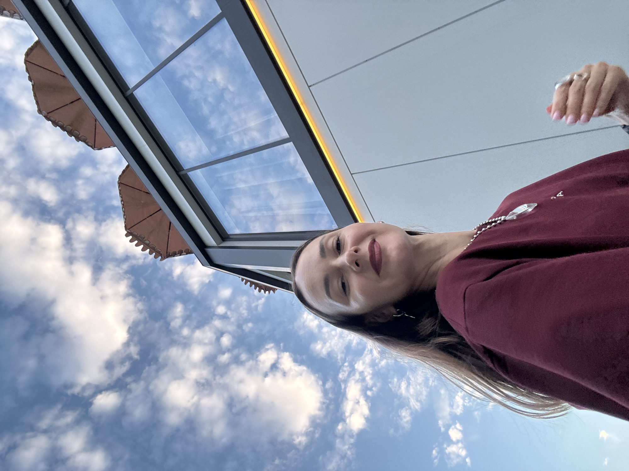 A low-angle, upward-facing selfie captures a woman in the foreground, looking directly at the camera with a slight smile. She has dark brown hair, styled loosely, and is wearing a maroon sweatshirt with a silver pendant necklace. Her right hand is raised, showing a ring on her index finger and manicured nails. Behind her, a modern building with large glass windows and an orange light strip is visible against a backdrop of a bright blue sky filled with scattered white clouds. The building's architecture suggests it could be a part of a hotel, restaurant, or commercial establishment. The overall impression is one of a casual, outdoor setting. While the exact location in Sochi is not specified, the architectural style and the clear sky are consistent with a city known for its coastal and mountainous landscapes, and resorts.