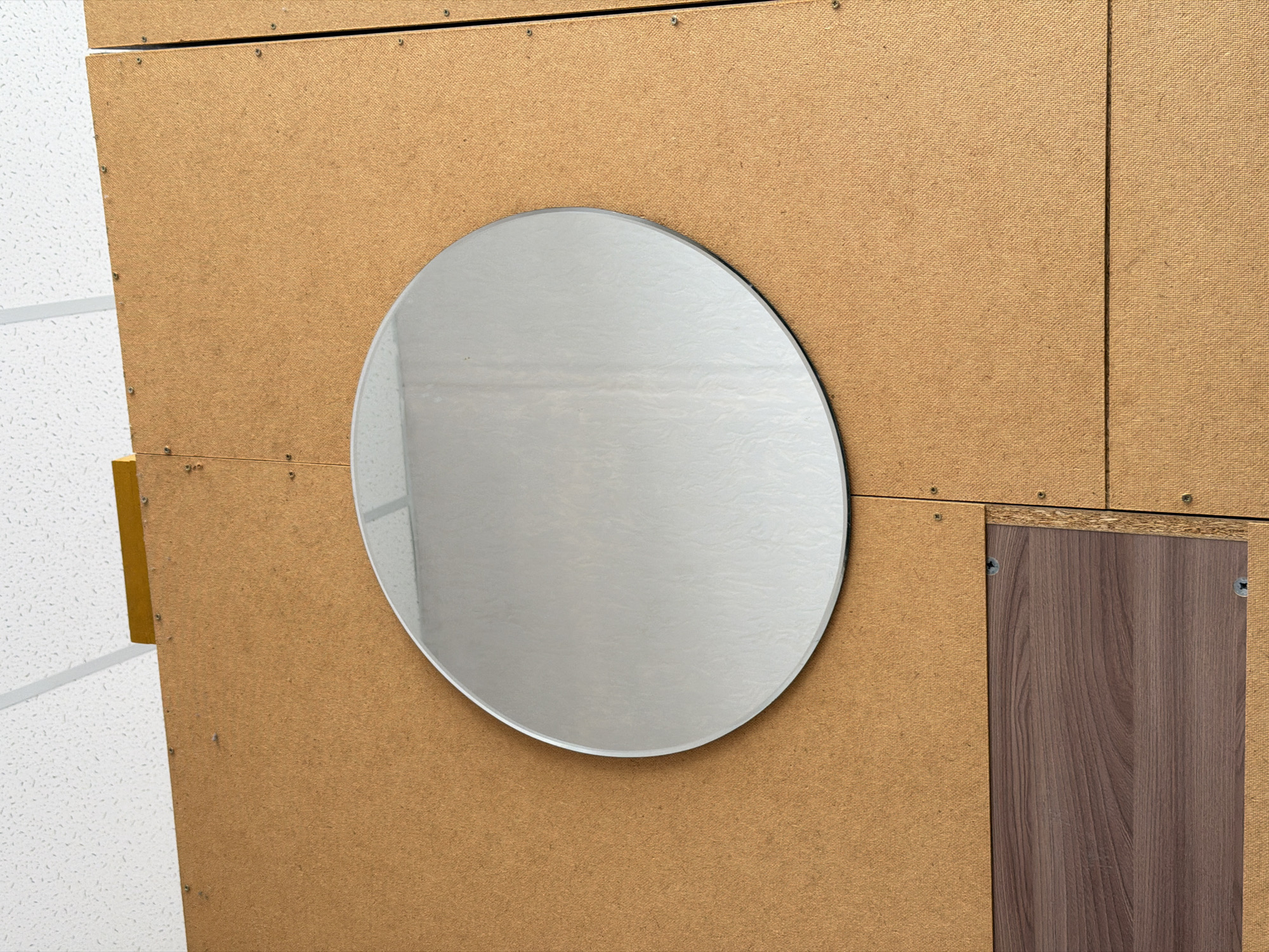 This media file presents a close-up view of an interior wall or partition, primarily constructed from light brown, coarse-textured particle board or oriented strand board (OSB). The panels are visibly joined with seams and secured by numerous silver-headed screws, particularly along the top and left edges.
A prominent feature is a large, irregularly shaped, oval-like mirror centrally placed against the particle board. The mirror's surface is highly reflective, showing a bright, indistinct light-toned interior, likely a ceiling or wall, with a subtle horizontal line visible within its reflection. The mirror appears to have a slightly beveled edge.
To the far left of the image, a portion of a white acoustic ceiling tile with a fine, speckled texture is visible, extending diagonally downwards. A vertical strip of bright yellow material, possibly a structural support or accent, is positioned between the ceiling tile and the particle board. In the lower right corner, a rectangular insert of dark brown wood-grain laminate or veneer is recessed into the particle board, secured by two visible screws.
The scene depicts a static interior detail, devoid of people or activity. The overall aesthetic suggests a functional, possibly industrial, institutional, or DIY environment. The lighting is bright and even, indicative of artificial indoor illumination. No text is visible in the image. The construction materials and methods point to a contemporary interior setting.