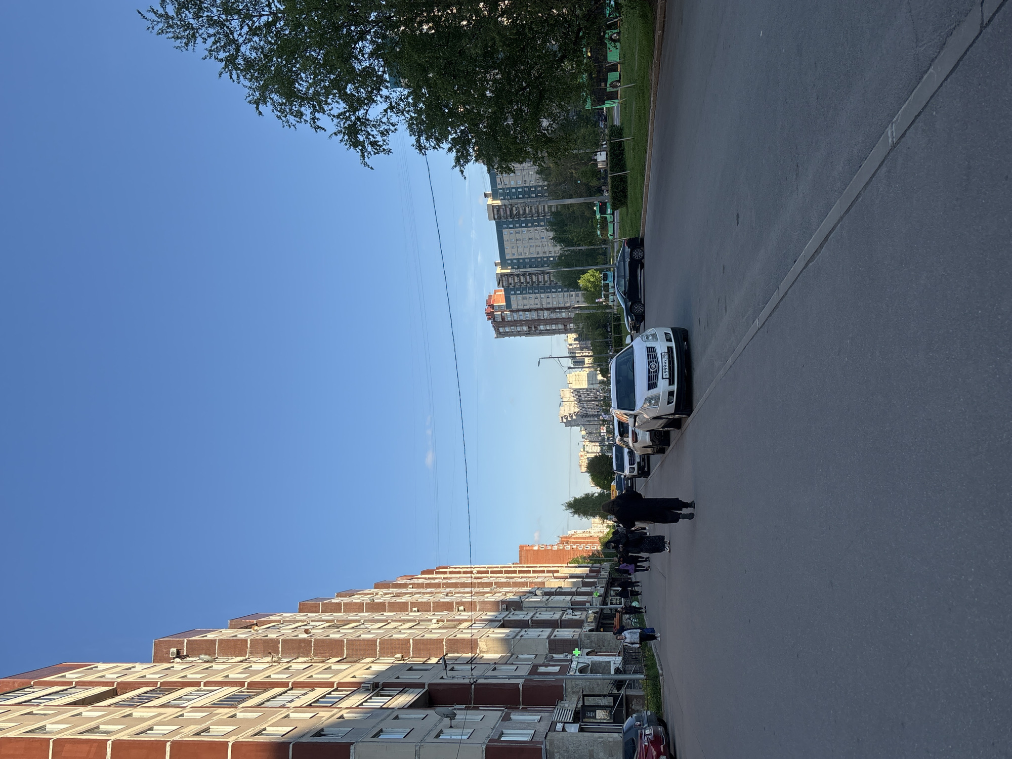 The image shows a street scene in Saint Petersburg, Russia. An apartment building lines the left side of the street, while the right side features parked cars and green spaces. Several pedestrians are walking on the sidewalk and along the road's edge. The sky is clear and blue. The cars include a white van in the middle of the road and a red car parked along the street. Overhead electrical lines are visible.