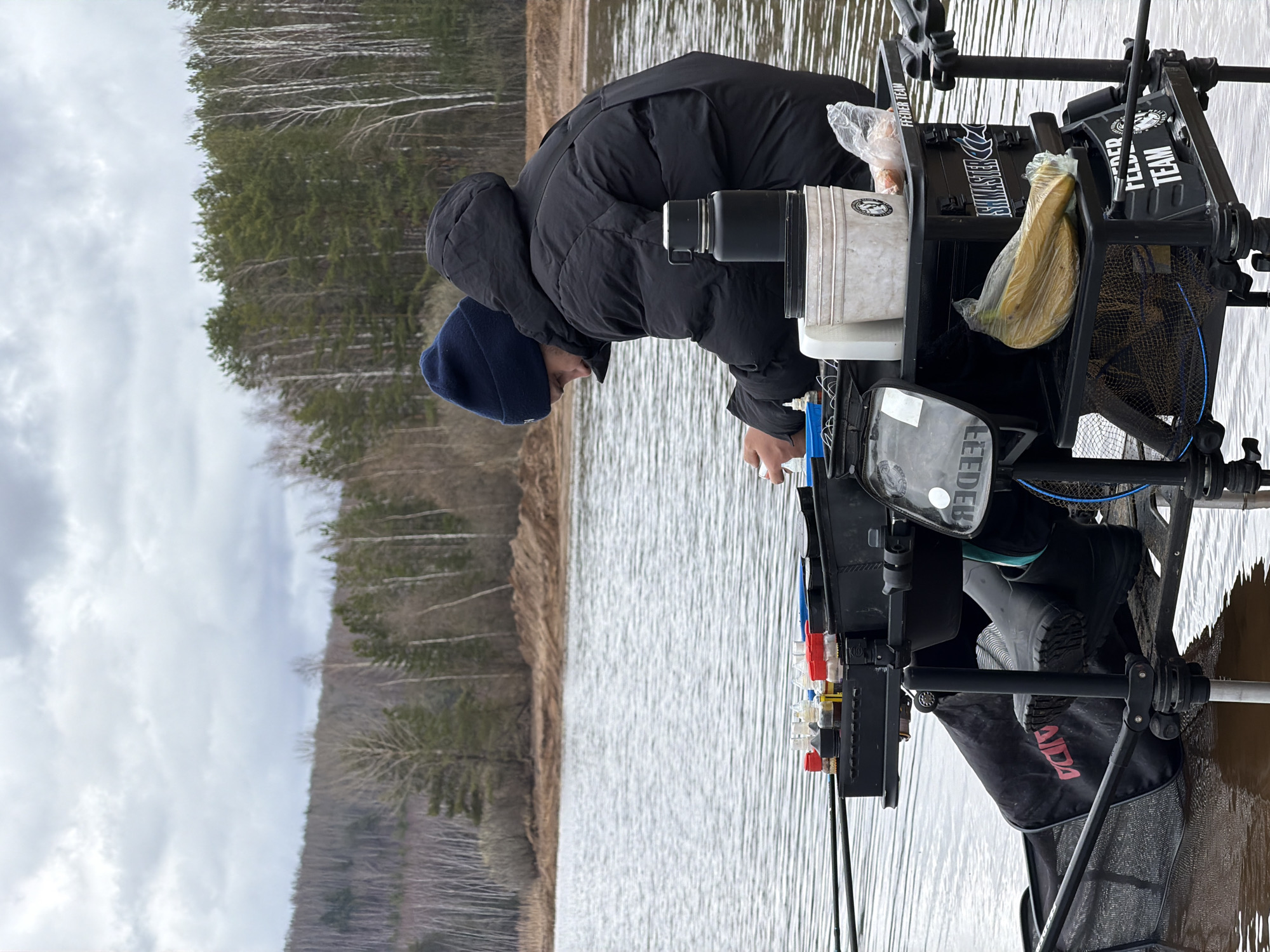 A man in a black puffer jacket and blue beanie is fishing on the shore of a lake. He is leaning over a black metal fishing platform with various fishing supplies. The platform is partially submerged in the water. Behind him is a view of a lake with trees on the opposite shore. The location is Chaikovsky, Russia.
