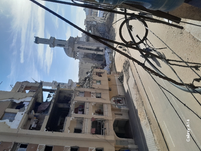 Extensive structural damage marks buildings, revealing exposed interiors and collapsed sections. A mosque with a prominent minaret stands amidst the destruction. Utility wires crisscross overhead, contrasting with the ravaged urban landscape below. A street runs alongside the damaged buildings. The sky is bright and partially clouded.