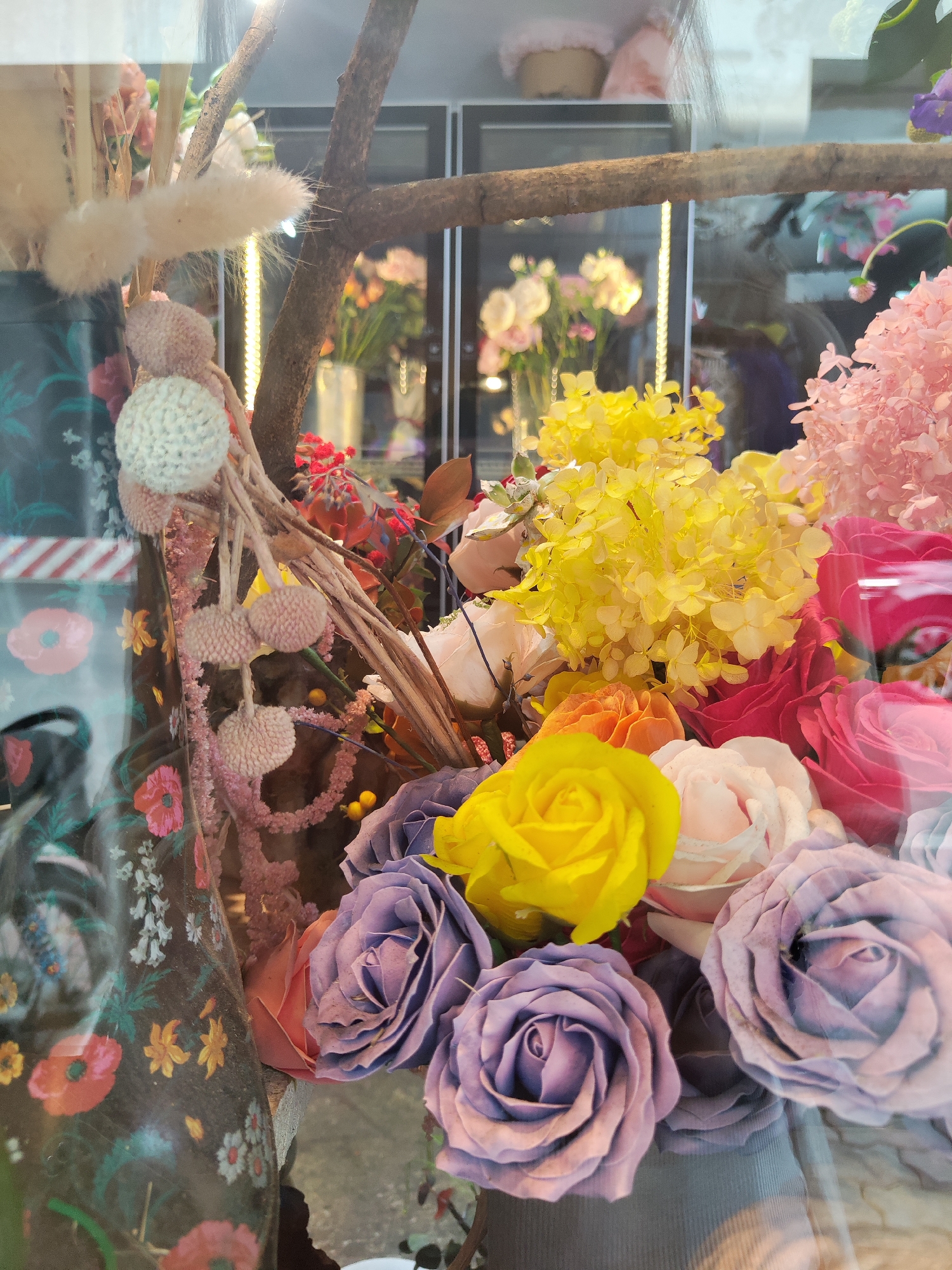 The image shows a floral arrangement in a display case, likely inside a shop. The foreground is dominated by colorful roses in various shades including lavender, yellow, red, orange, and white, interspersed with yellow and pink hydrangeas. A decorative branch crosses the top of the frame. To the left, a vase with a floral pattern can be seen. Knitted pom-poms and other decorative elements are arranged around the main flower display. The display is behind glass and reflects the outside street scene. In the background, inside of the flower shop, there is a variety of other flowers in vases with glowing string lights. Based on available information, it could potentially be in Thủ Đức, Vietnam, but there is no specific evidence in the picture to confirm this location.