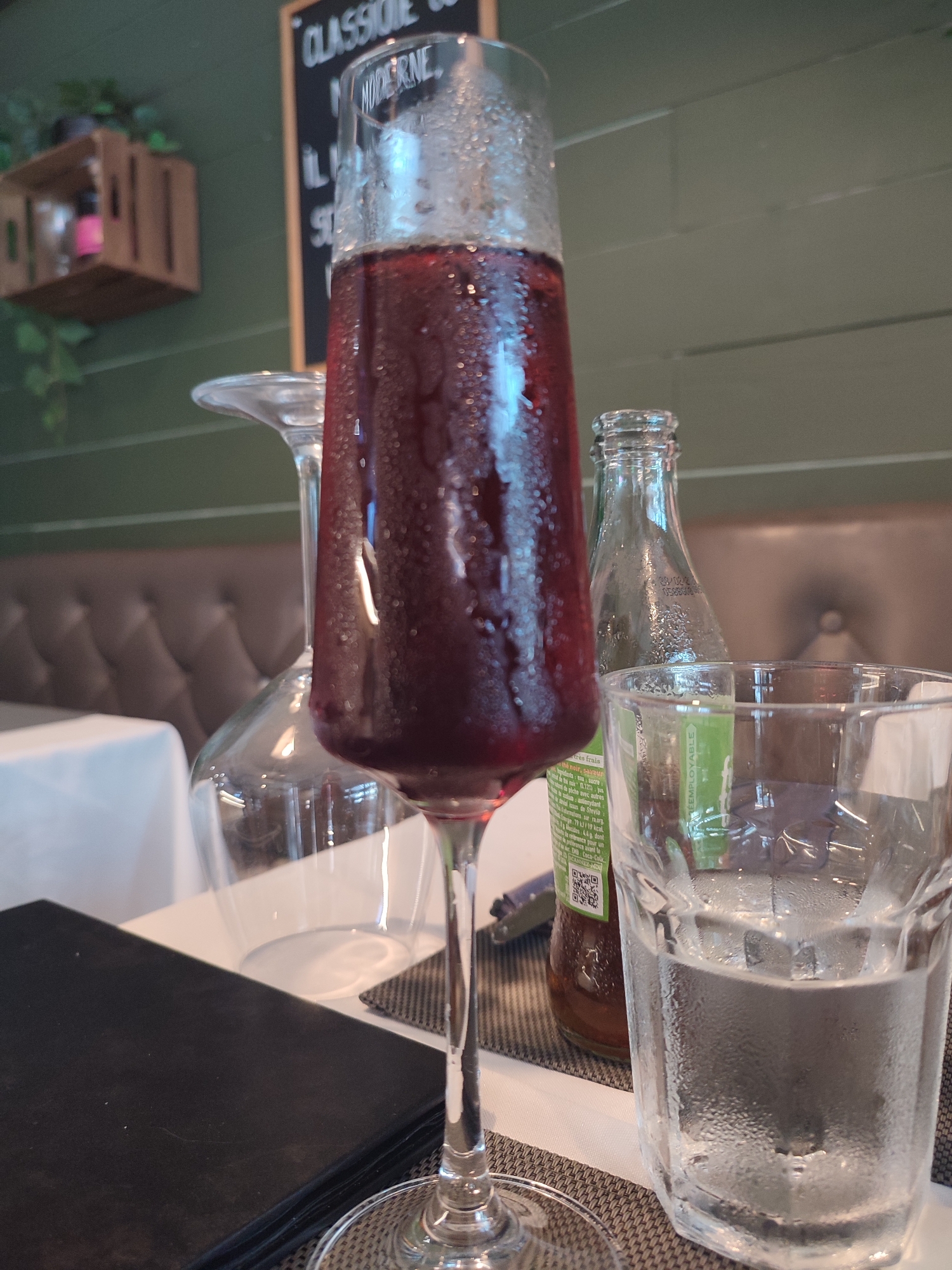 A table setting at a restaurant in Hyères, France features a tall, narrow glass filled with a dark red liquid, likely wine or a cocktail, and condensation on its exterior. Beside it, a clear glass contains water with condensation. A clear glass bottle, possibly containing a beverage, stands behind the drinks. A dark-colored menu lies on the table in the foreground. In the background, a brown tufted booth and green walls are visible. A small wooden crate with plants is mounted on the wall. A blackboard with French text is also present in the background.