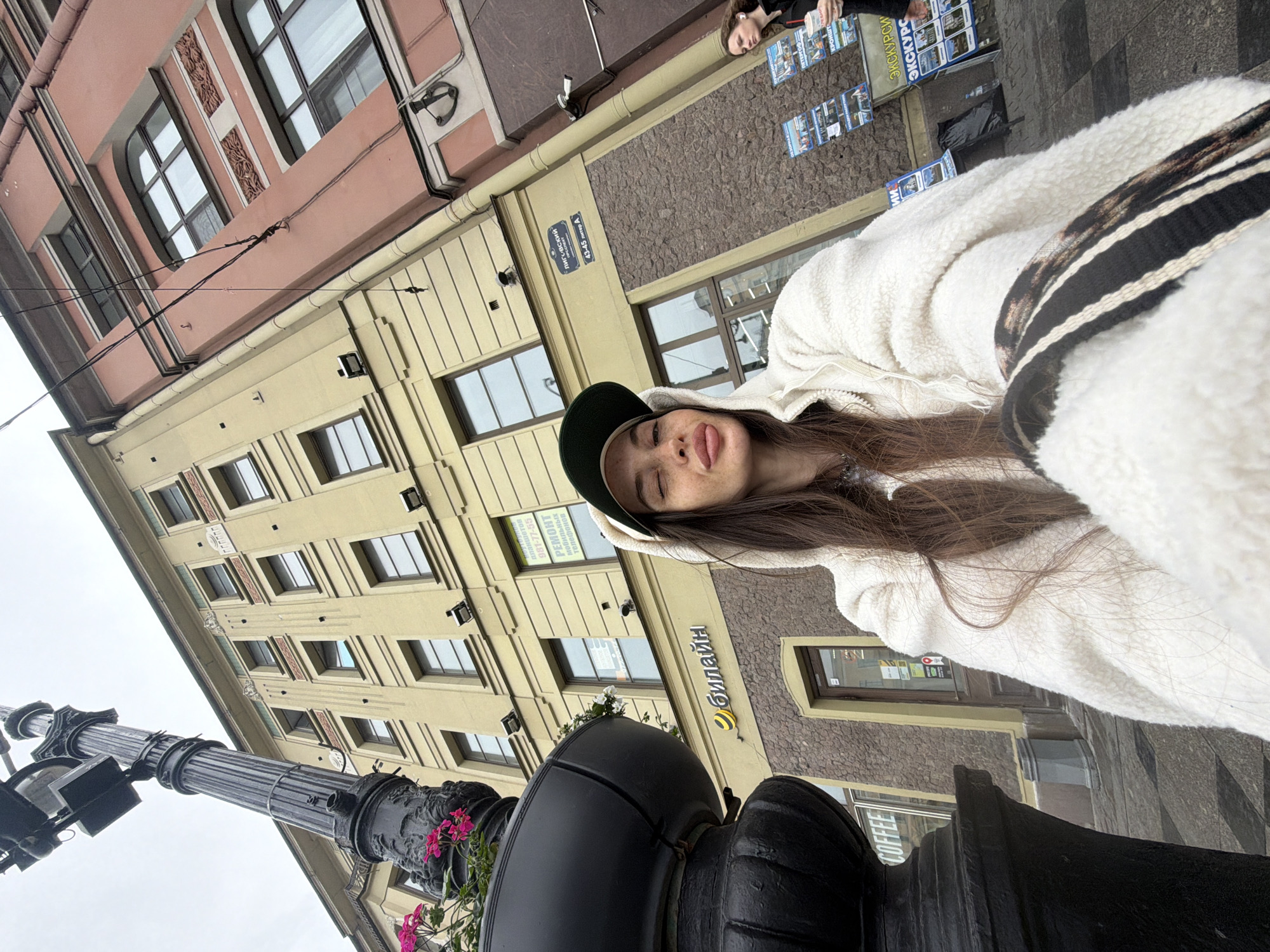 The image depicts a young woman taking a selfie outdoors in Saint Petersburg, Russia. She is wearing a white hooded jacket and a baseball cap, and her face is visible as she winks. Behind her, two buildings are visible; one is yellow with arched windows and decorative elements, and the other is pink with similar architectural features. A street lamp with floral decorations stands to the left of the frame. Signs for "Beeline" and "COFFEE" are visible on the yellow building, along with Russian text indicating "43-45 LIGOVskiy PROSPEKT". In the background, two other individuals are walking, one of whom is holding a cup and looking towards the camera. The sky is overcast.