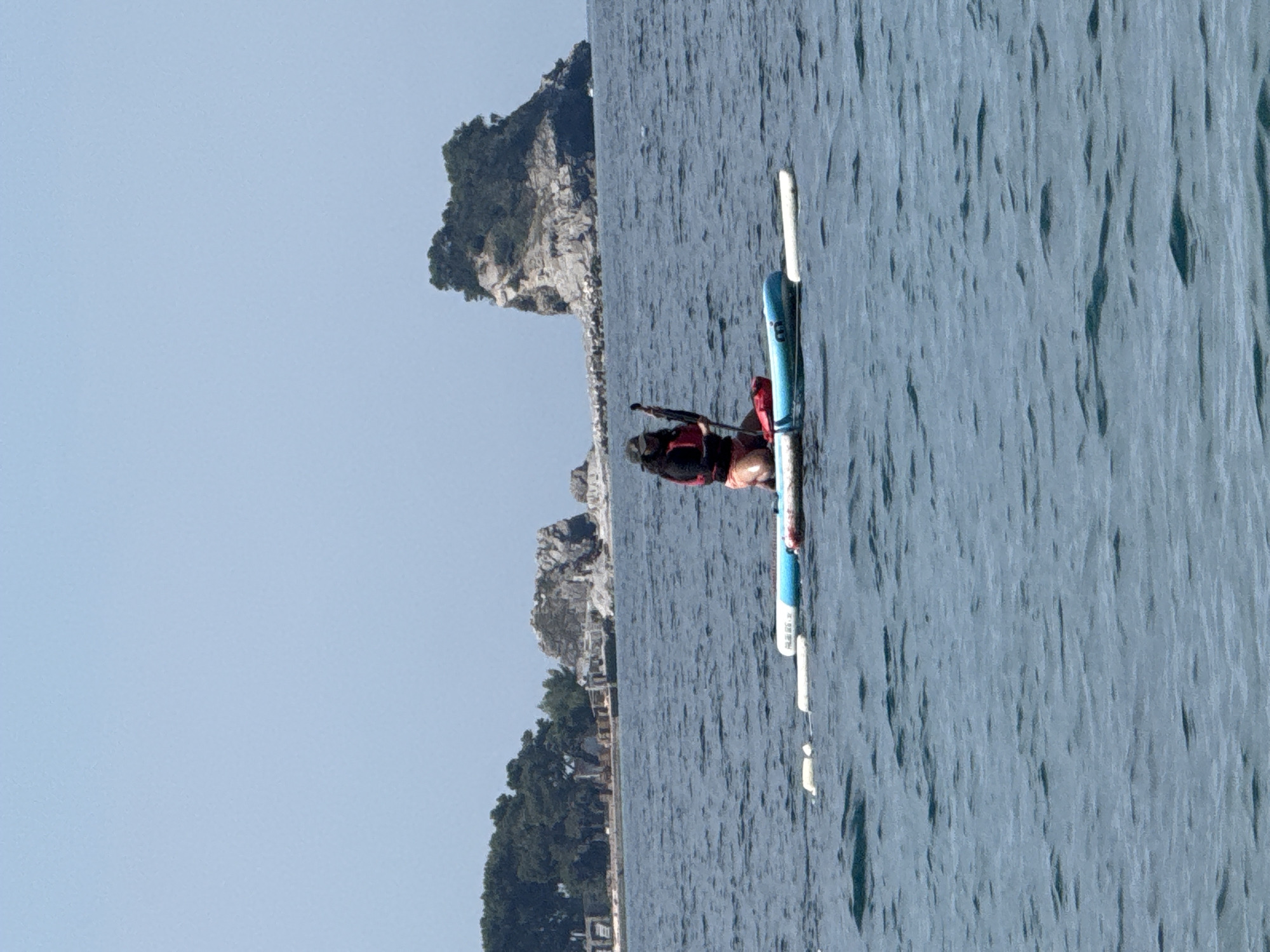 A medium outdoor shot shows a person on a paddleboard in the sea near Budva, Montenegro. The person, wearing a red life vest, is paddling. The sea is slightly choppy. The coastline, featuring trees and rock formations, is visible in the background. The sky is light blue.