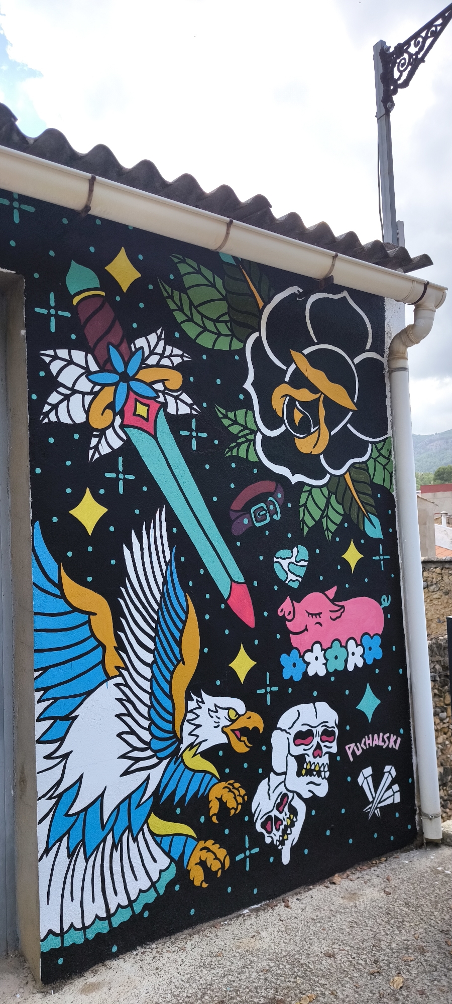 This image shows a vibrant mural painted on the exterior wall of a building in Fanzara, Spain. The mural, which occupies a significant portion of the wall, features a dark background with various illustrative elements in bright, contrasting colors.
Dominating the left side of the mural is a depiction of a stylized eagle in flight, rendered in white, blue, and yellow with black outlines. Its wings are spread wide, and its talons are extended. To its right, a turquoise and red dagger is positioned diagonally, with a blue flower and a yellow diamond at its hilt, surrounded by white floral or leafy elements. Above the dagger, a black rose with white petals and a yellow center is depicted, with green leaves extending downwards.
In the upper right quadrant, a pink pig with closed eyes is shown resting on a bed of blue and white flowers. Below the pig, two white skulls with red eyes and teeth are painted, one slightly overlapping the other. To the right of the skulls, the name "PUCHALSKI" is written in pink lettering, and below it, three crossed nails are depicted in white. Scattered across the black background are small blue dots, teal crosses, and yellow stars.
The building's exterior features a grey-tiled roof with a white gutter running along the edge, partially visible at the top of the image. A portion of a black street lamp with decorative metalwork is visible in the upper right corner. The lower part of the image shows a grey concrete ground surface, with scattered dry leaves. In the background, beyond the building, hints of trees and distant hills can be seen, along with a cloudy sky.