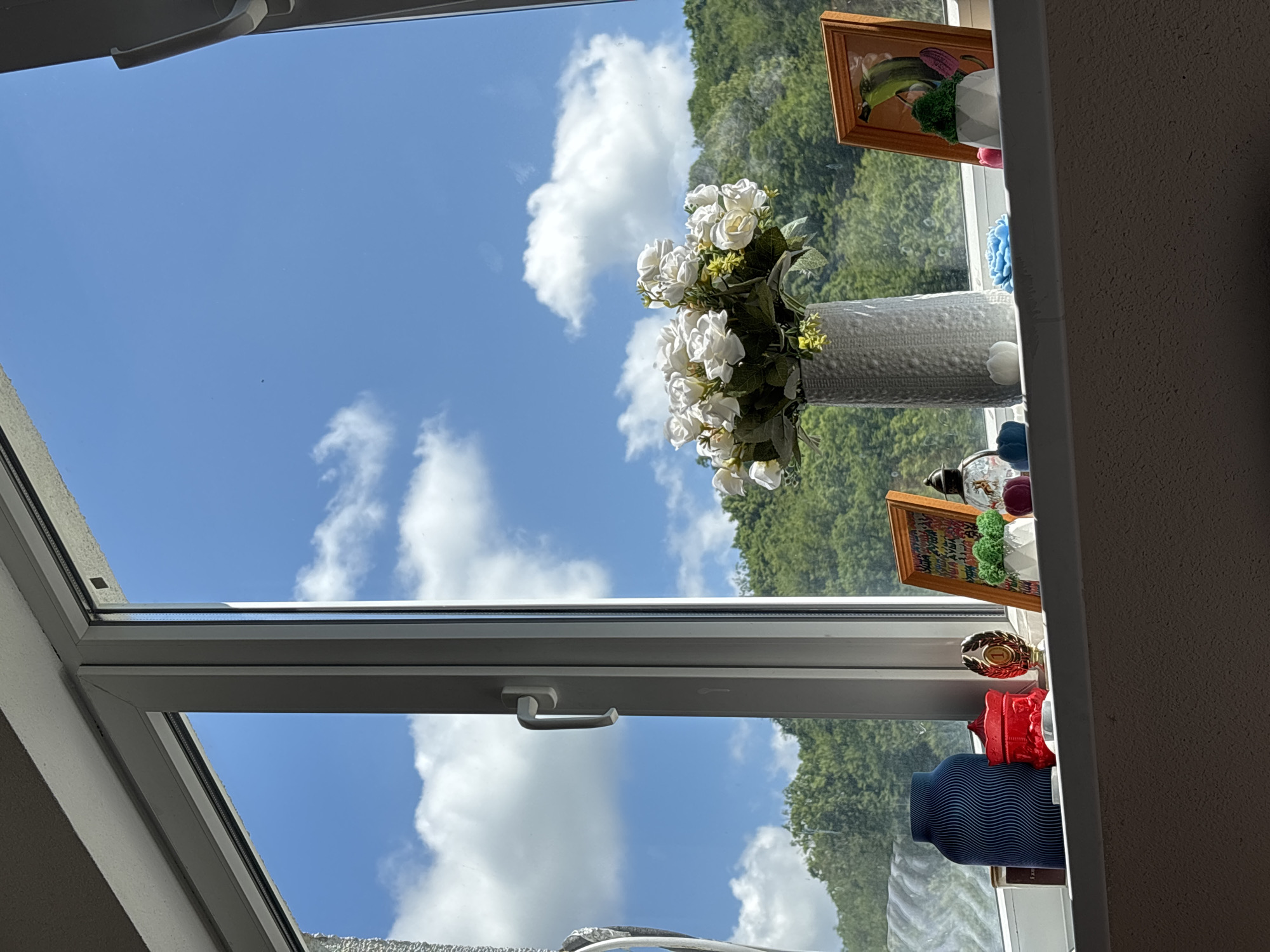 The image shows a window with a view of a green, tree-covered hillside under a blue sky with scattered white clouds. On the windowsill, there are several decorative objects, including a vase of white artificial flowers, a blue vase with a wavy pattern, a red lidded container, a trophy, a framed picture of a green object resembling a cucumber, and several small, colorful decorative items including what appear to be small potted plants and candles. The window itself is white and has a handle on the right side. The scene suggests an indoor setting looking out onto a natural landscape. The specific location in the city of Sochi, Russia, cannot be determined from the image alone.